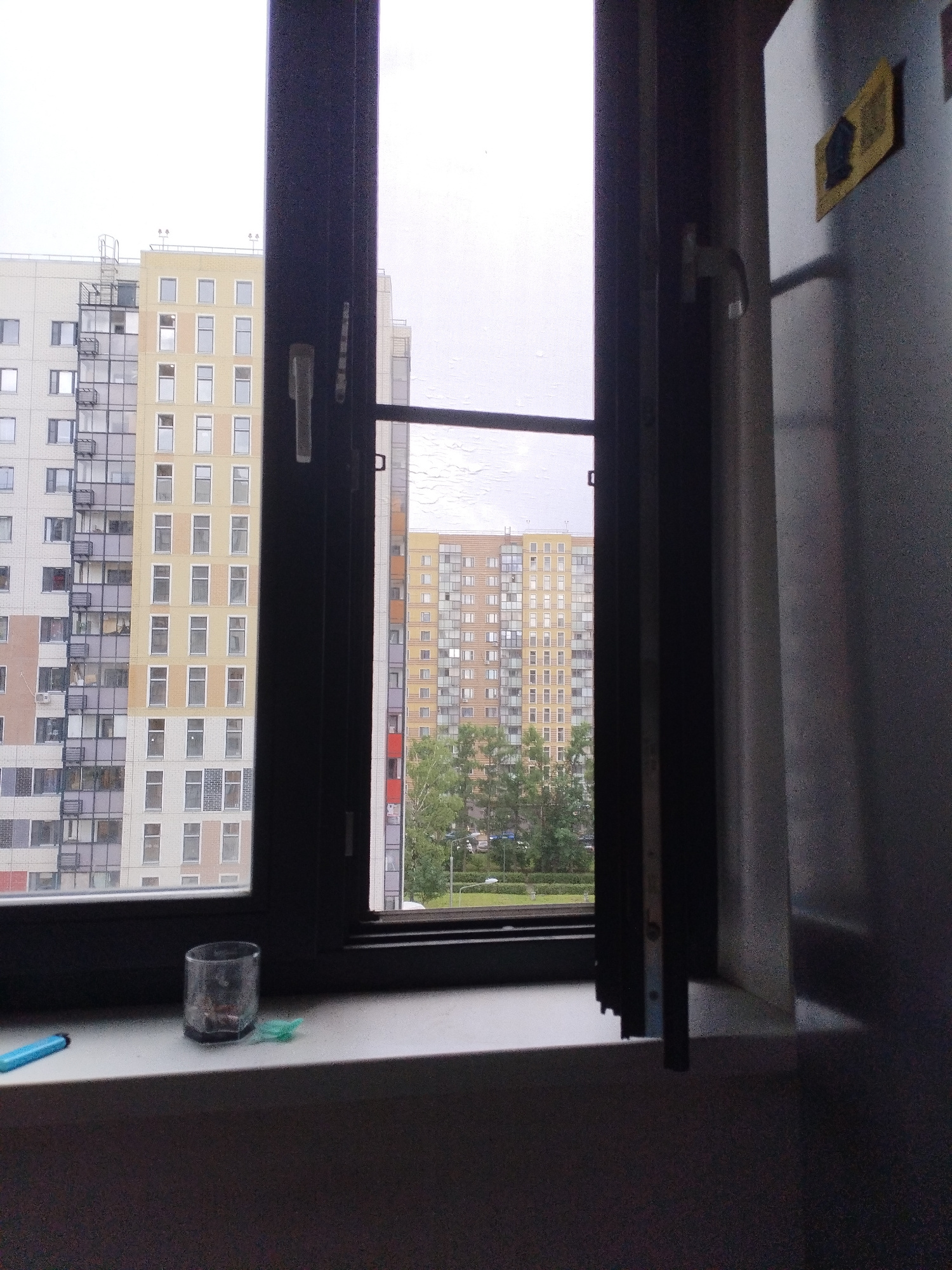 A medium shot captures a view through a partially open window, looking out at two multi-story apartment buildings. The building on the left is predominantly light grey and white with a darker grey section featuring numerous balconies. The building in the center is a lighter yellow-orange with many windows. The building on the right is also a lighter color with rows of windows and a visible green tree in front of it. The sky is overcast and white. On the windowsill in the foreground, a clear glass, a blue pen-like object, and a small green item are visible. To the right of the window, a large silver appliance, possibly a refrigerator, is present, with a yellow square sticker on its side. The scene appears to be an urban environment.