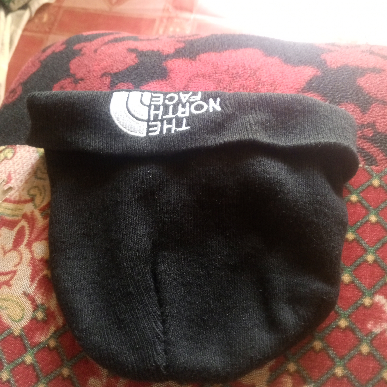 A black knitted beanie with a white embroidered logo is laid flat on a patterned surface. The logo reads "THE NORTH FACE" and appears on the cuff of the beanie. The beanie is folded in half. The patterned surface is a rug with a red and black camouflage pattern and a red, green, and gold geometric pattern. The lighting suggests an indoor environment.