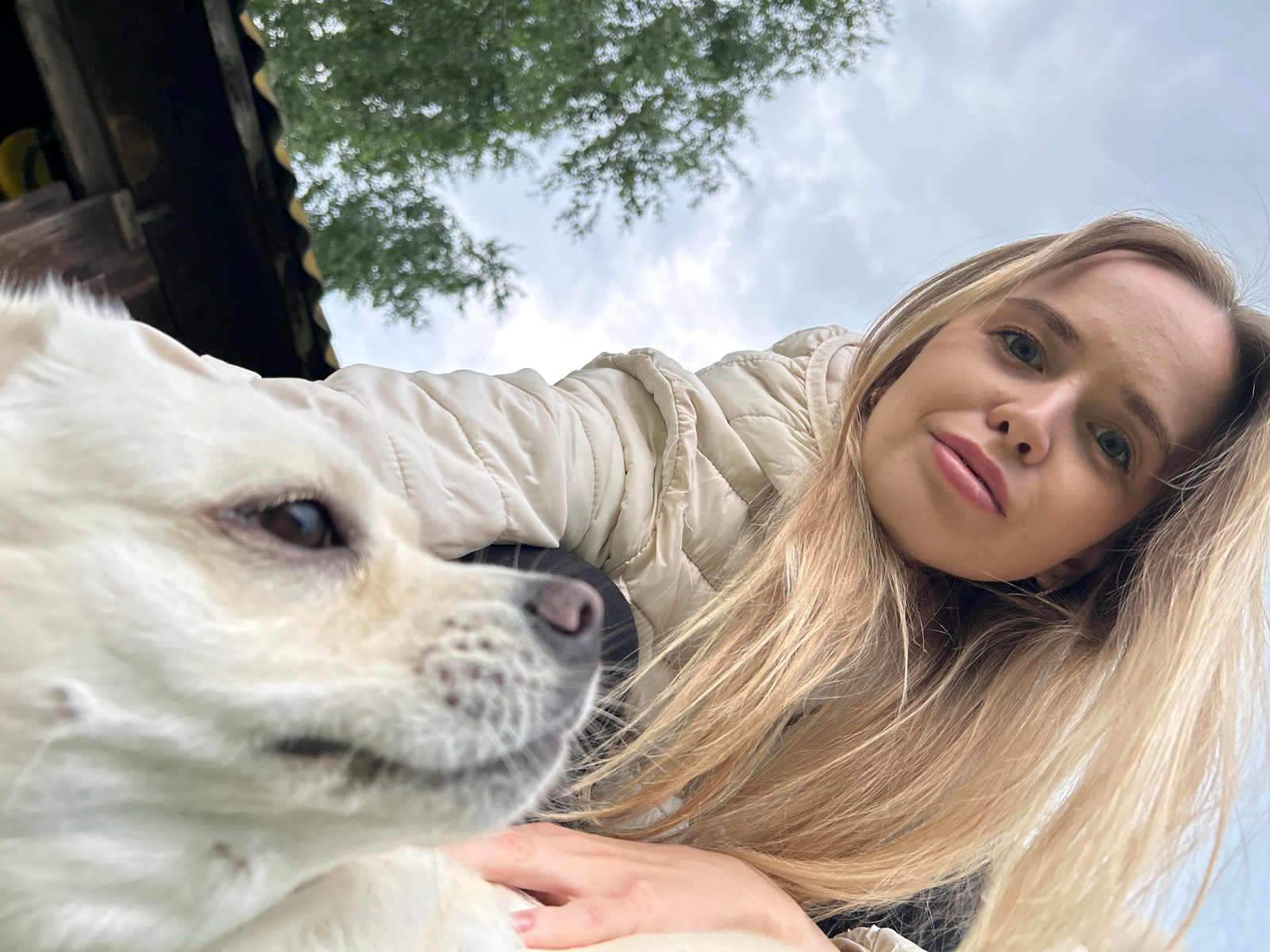 A young blonde woman is taking a selfie outdoors with a white dog in the foreground. The woman is wearing a cream-colored puffer jacket and her hair is falling around her shoulders. The dog, which appears to be a Pomeranian or similar breed, is looking to the left of the frame, with its face partially out of focus. Behind them, a cloudy sky and green foliage are visible, along with part of a dark-colored structure, possibly a roof. The location is not specified in the image.