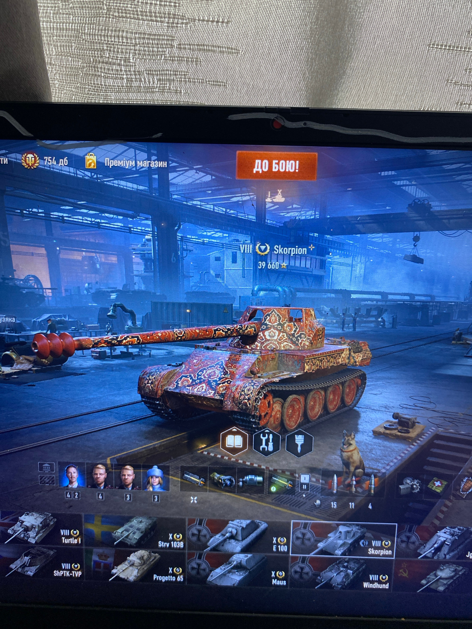 The image depicts a scene from a video game interface, specifically the garage or hangar screen of a tank simulation game, likely World of Tanks. The user is viewing a heavily decorated tank with a red, gold, and blue ornate pattern. The game interface displays various elements: a prominent "GO TO BATTLE!" button in orange, the tank's designation "VIII Skorpion" with associated stats, and a selection of other tanks available to the player. Crew members are also visible in the interface, represented by portraits with numbers indicating their rank or experience. The background appears to be a stylized industrial environment, likely representing a game asset and not a real-world location. There is no indication or possibility of this image being related to Szczecin, Poland.