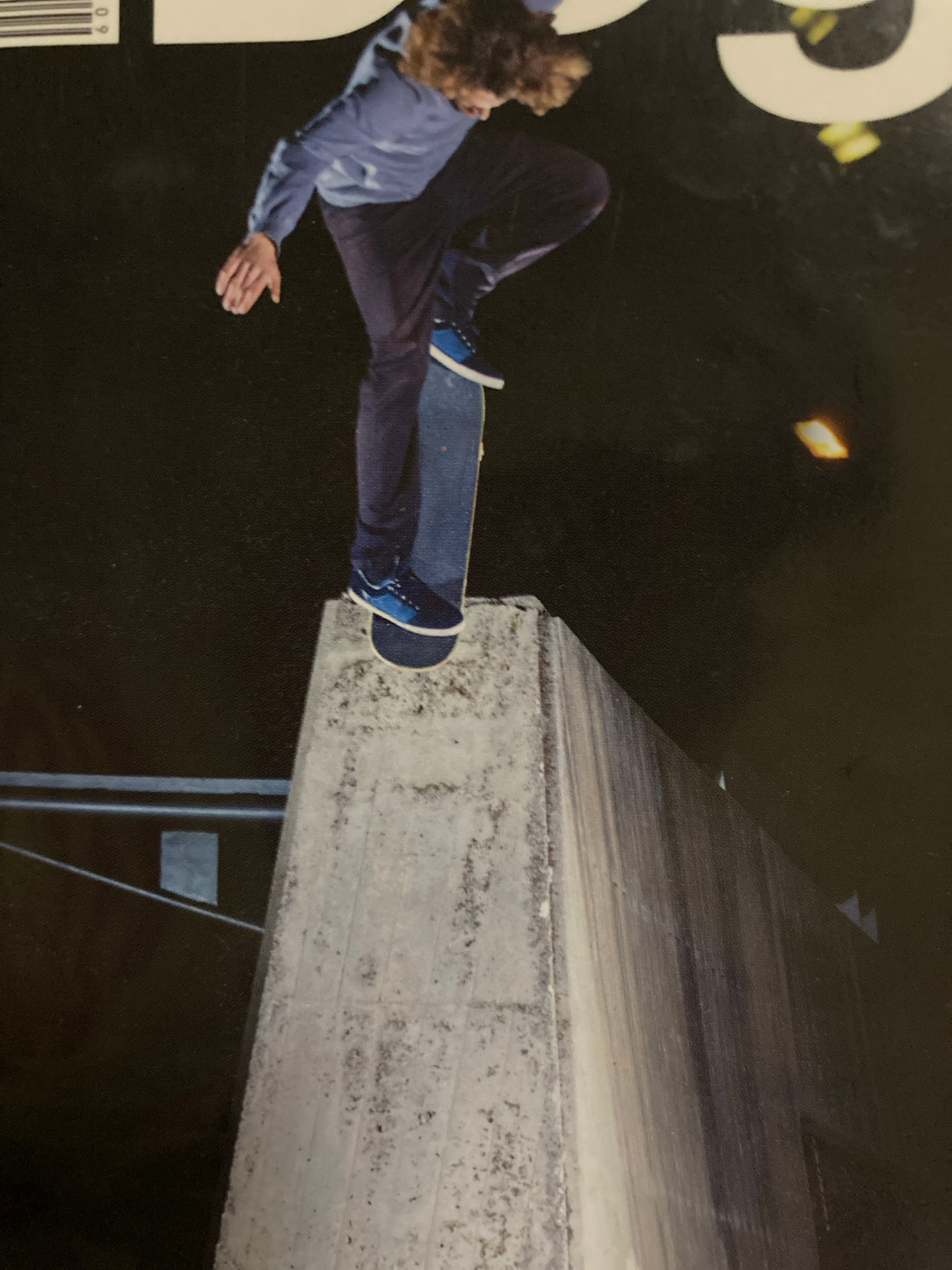 The image shows a skateboarder performing a trick over a concrete structure, likely a bridge or overpass. The skateboarder, dressed in dark pants and a blue shirt, is captured mid-air with one leg extended. Their skateboard is beneath their feet, also in motion. The background is dark, suggesting it is nighttime or a very dimly lit environment. A bright, rectangular light source is visible in the upper left quadrant of the image, adding a dramatic element. The concrete structure appears rough and textured, with some signs of wear and tear. The overall scene conveys a sense of action, athleticism, and perhaps a touch of danger due to the height and the dark surroundings. The skateboarder's body language suggests focus and skill. There is some text visible in the bottom left corner, possibly a barcode and numbers, suggesting this image may be from a magazine cover or advertisement. The location context of Sabadell, Spain, doesn't provide specific visual cues in the image itself, but it places the urban setting of the skateboarder's activity within that Spanish city.