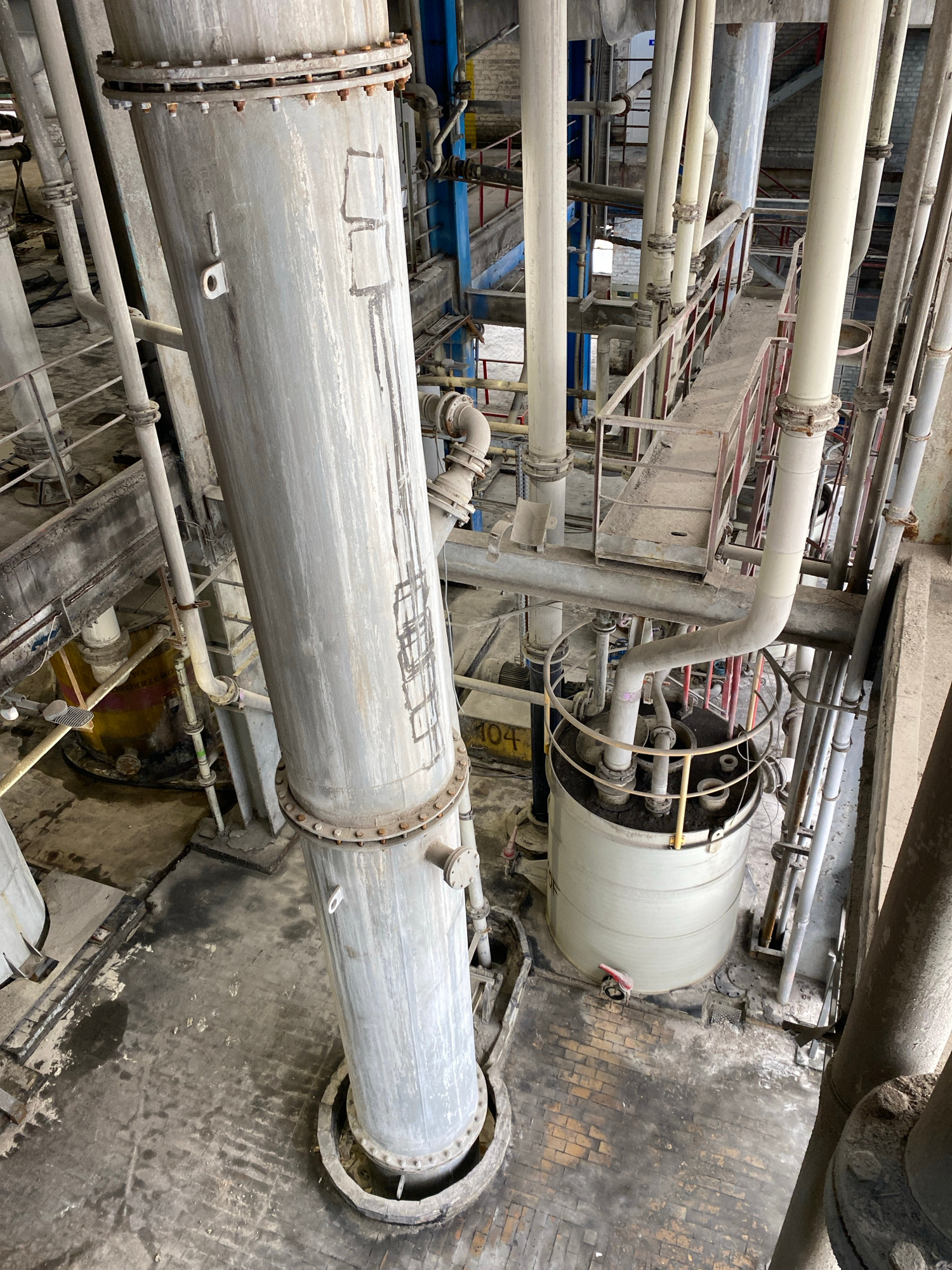 This image depicts the interior of an industrial facility, likely a processing plant, featuring large metal pipes, cylindrical tanks, and elevated walkways. The scene is characterized by industrial machinery, with various interconnected pipes of different diameters dominating the frame. The environment appears dusty and somewhat dilapidated, with exposed brickwork and concrete flooring showing signs of wear and discoloration. In the background, a multi-level structure with railings and stairs is visible, suggesting an organized system for maintenance or operation. The lighting is artificial, creating a utilitarian ambiance typical of such industrial settings. There is no indication of human presence in the image. The presence of signage or equipment labels in Polish might suggest the location is Police, Poland, a town known for its industrial zones. The overall impression is one of heavy industrial activity and infrastructure.