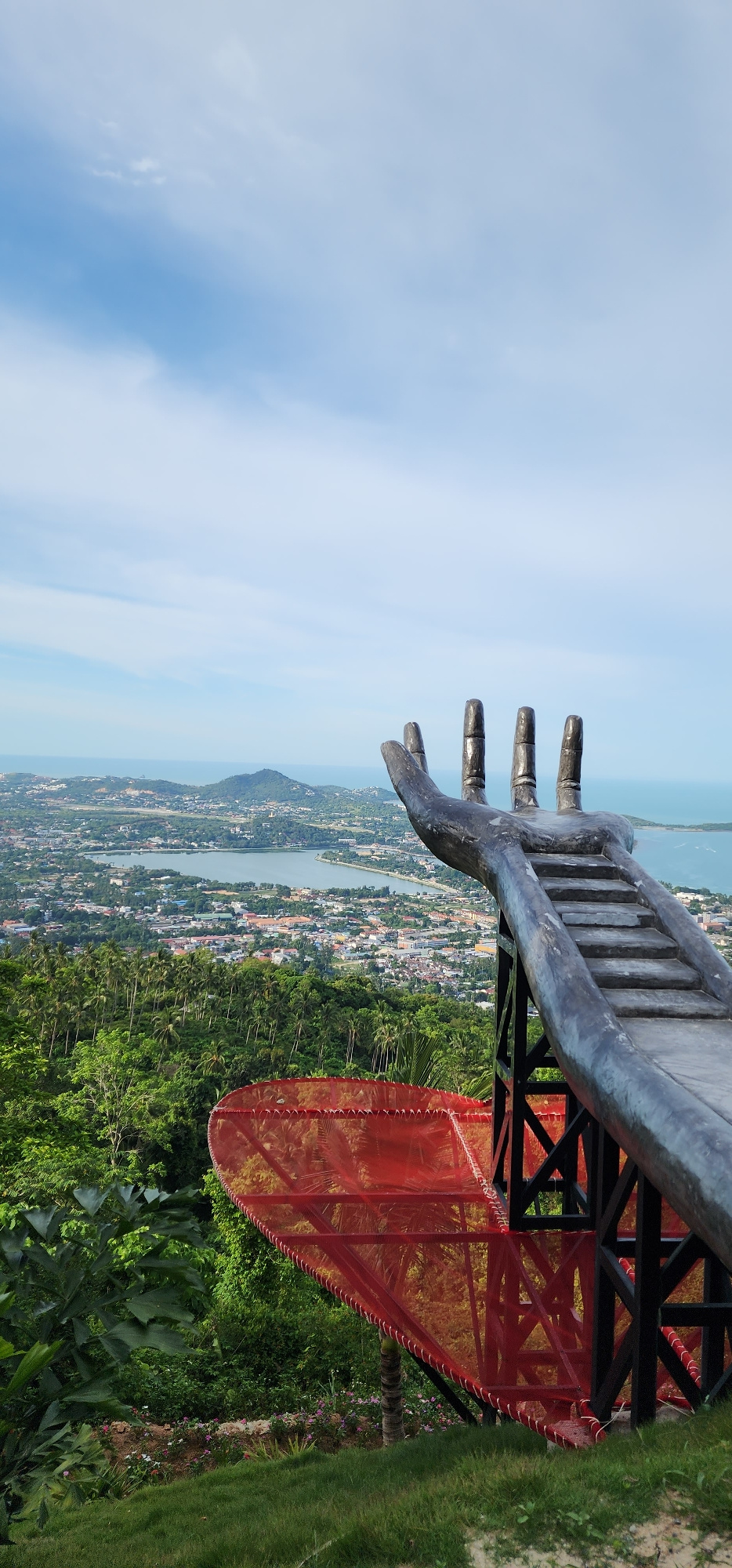 This vertical, eye-level, medium shot captures a view from a scenic overlook on Ko Samui, Thailand. In the foreground, a large, dark-colored, stylized sculpture of a hand extends into the frame from the right, with stairs integrated into the palm and fingers, appearing to lead upwards. Below the hand sculpture, a striking red mesh structure, triangular in shape and supported by black metal beams, juts out. The background reveals a panoramic vista of a coastal city with a lake at its center, nestled between lush green hills and the turquoise ocean. The sky above is a clear blue with scattered white clouds. The overall impression is of an elevated vantage point offering a breathtaking landscape view.