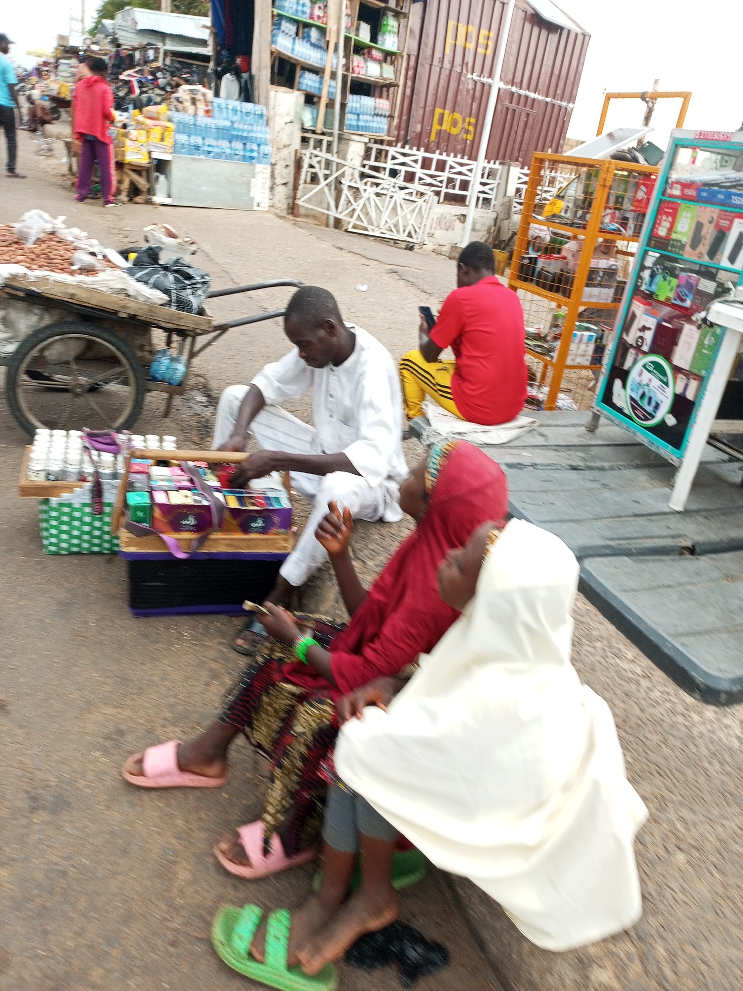 In Gombe, Nigeria, at what appears to be a roadside market, a man in a white garment sits on the ground next to a wooden cart filled with various products. He is attending to a colorful display of small boxes and jars arranged on a smaller wooden platform. To his right, a person wearing a red shirt and yellow pants is seated, looking at their mobile phone. In the foreground, two individuals, one wearing a red headscarf and the other a white head covering, are also seated. The person with the red headscarf is dressed in a patterned fabric, and both are barefoot. Behind the seated individuals, a large cart with a wheel holds bags and other items. In the background, stalls and shelves are visible, stocked with goods like bottled water and electronic devices. The overall scene depicts a bustling marketplace with vendors and customers engaged in commerce.