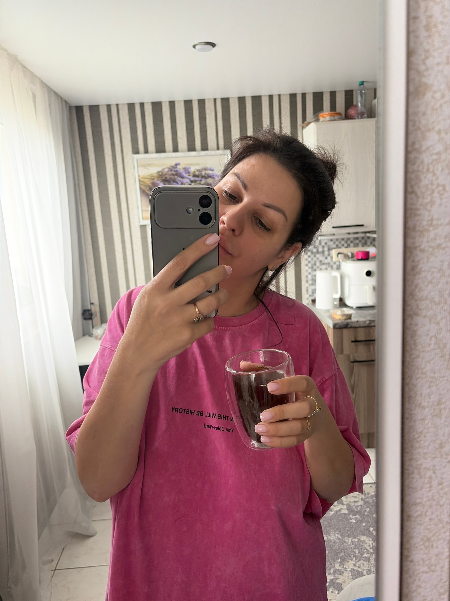 A woman with dark hair tied back in a bun is taking a mirror selfie in what appears to be a kitchen. She is wearing a bright pink t-shirt and holding a clear glass filled with a dark liquid, possibly coffee, in her right hand. Her left hand is holding a smartphone, capturing the reflection. She is also wearing two rings on her left hand. The background shows striped wallpaper, a framed picture of lavender on the wall, and kitchen cabinets with appliances. The presence of a water bottle on a shelf suggests a domestic setting. The location is not specified in the image or context provided.