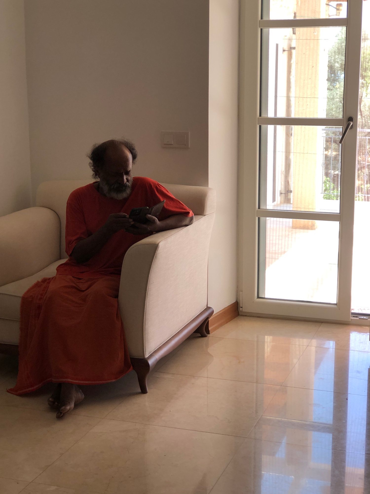 A man in an orange robe sits on a cream-colored armchair, looking down at his phone. His feet, bare and dark-skinned, are visible beneath the hem of his robe. The armchair is positioned on a tiled floor reflecting the bright light from a large glass door and window on the right side of the frame. The door and window offer a view of an outdoor space with a railing and foliage. The room is sparsely furnished, with a light-colored wall on the left. The scene appears to be indoors, possibly in a residential setting in Kaş, Turkey, suggested by the architecture and lighting characteristic of the region.