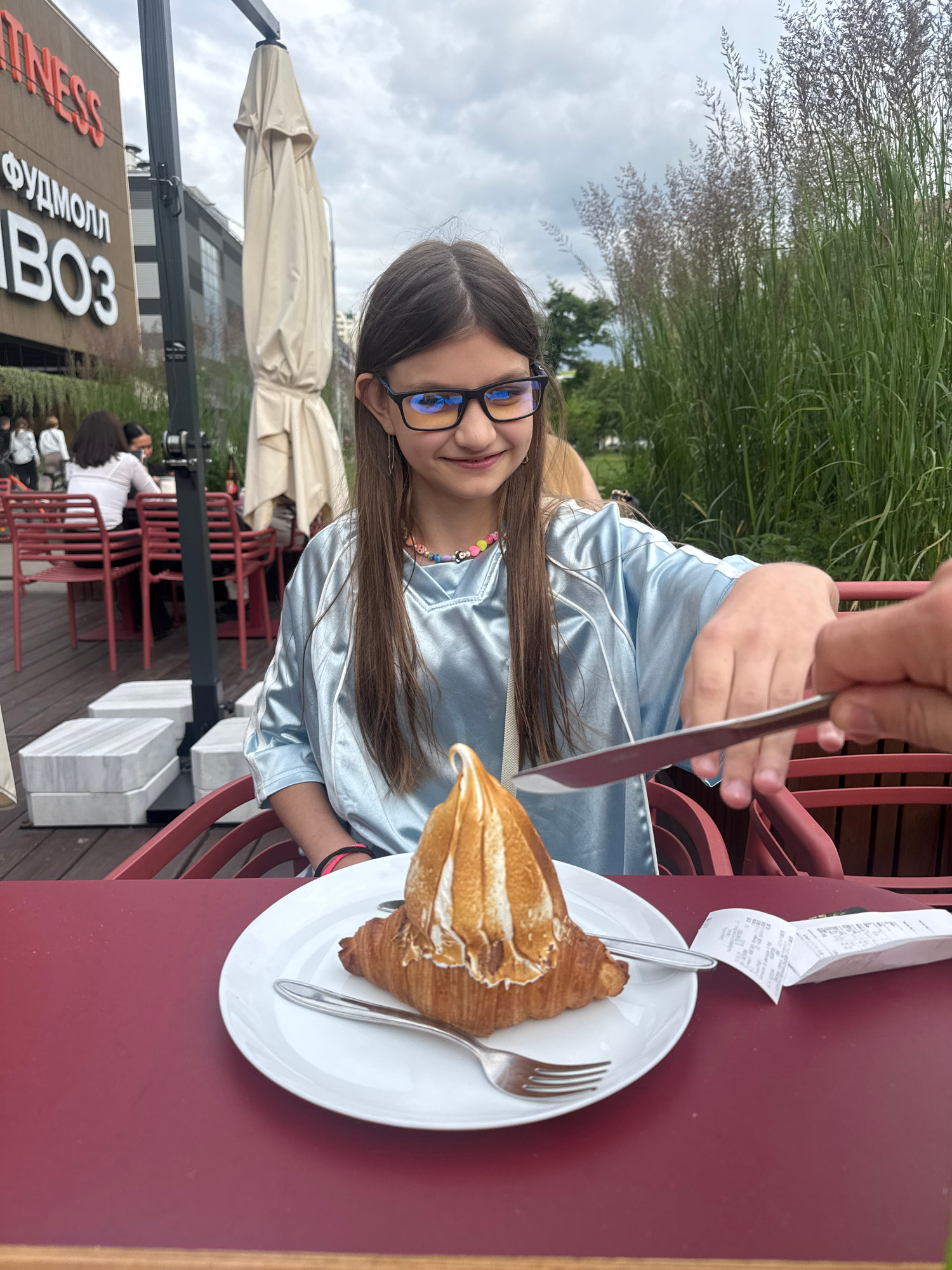 In this vertically oriented image, a young girl wearing glasses and a light blue, silky outfit is seated at a red table outdoors. She is smiling and looking towards the camera. Her hair is long and brown. A croissant topped with a meringue-like substance sits on a white plate in front of her. A fork and knife are also present on the plate. A hand, presumably of another person, is partially visible at the top of the frame, holding the knife as if to cut the pastry.
In the background, tall grasses are swaying, and a cloudy sky is visible. To the left, there's a partial view of a building with signage that includes Cyrillic text. One prominent sign reads "ФУДМОЛЛ" and "ВОЗ". The scene appears to be set in a cafe or restaurant with outdoor seating, indicated by the red chairs and tables. The overall mood seems relaxed and pleasant, possibly during a casual meal. The time of day appears to be daytime, given the natural light.