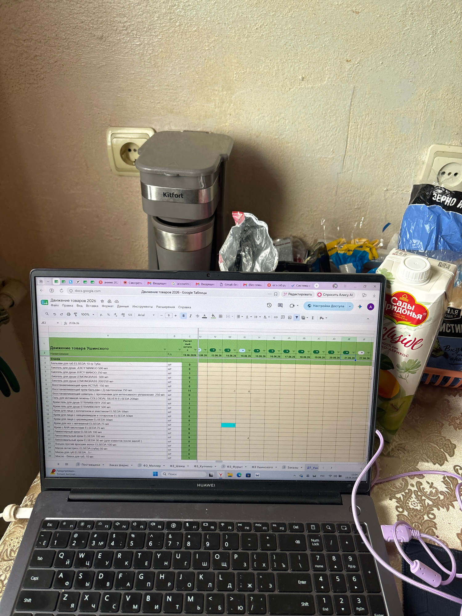 The image shows a laptop open and displaying a spreadsheet titled "Движение товара 2026" (Goods Movement 2026). The spreadsheet lists various cosmetic and personal care items from the brand "Elsedа" and their quantities. The laptop is placed on a table in an indoor environment, likely a home setting. To the left of the laptop, there is a Kitfort coffee maker. To the right, there is a carton of juice and some other items, including plastic bags. The background appears to be a textured wall with an electrical outlet. The overall scene suggests a workspace where someone is managing inventory or sales data.