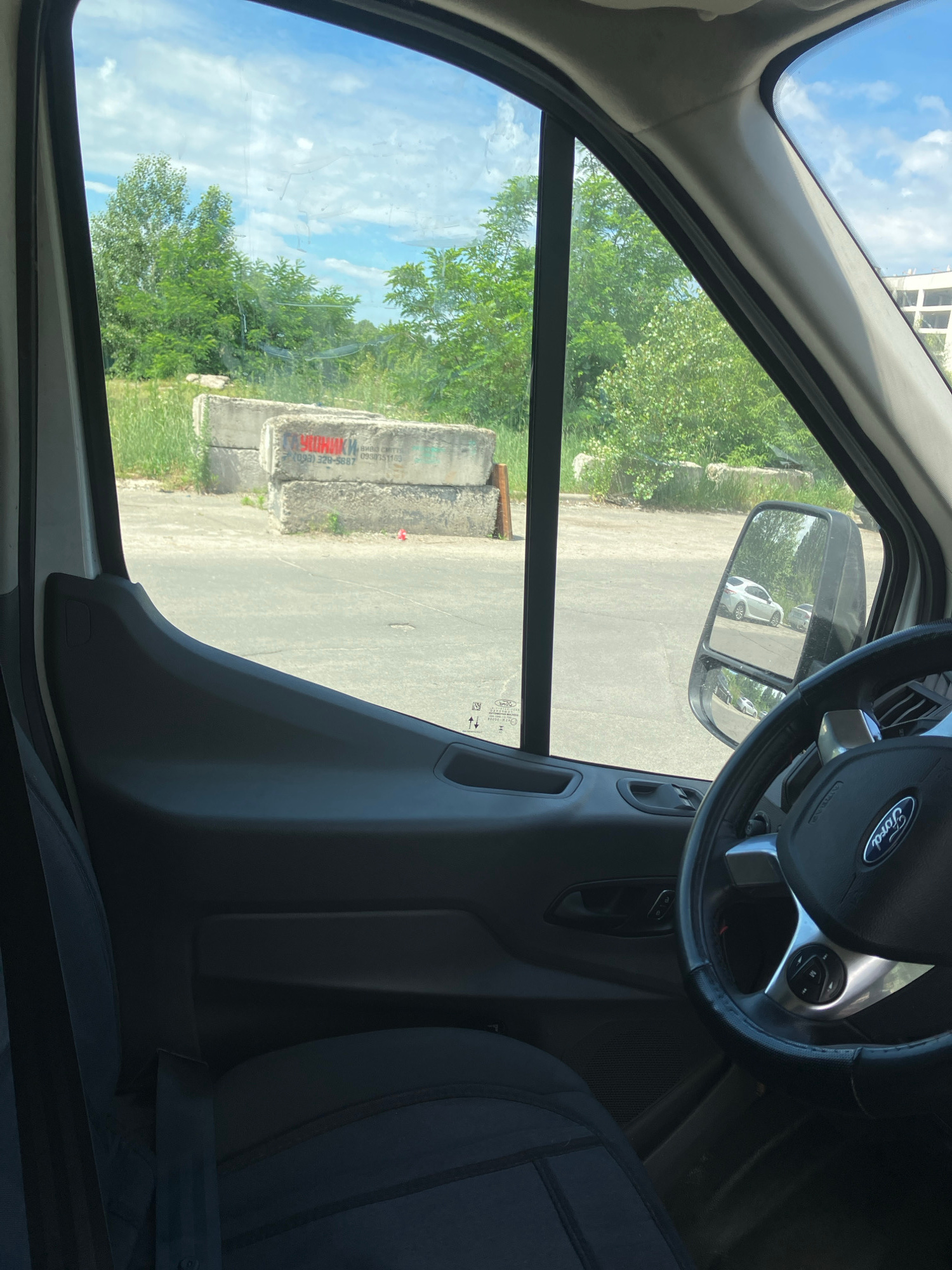 The image shows a view from inside a white Ford van, looking out the driver's side window. The van's interior is visible, including the dashboard, steering wheel with a Ford logo, a portion of the driver's seat, and the door panel. Outside the window, concrete blocks are stacked on the ground, with red graffiti on them. The graffiti appears to say "ЧОРОНИКИ" and a phone number, "(093) 328-5887". Beyond the blocks, there are trees and a cloudy blue sky. In the side mirror, two cars are visible parked on an asphalt surface. The scene appears to be an outdoor area, possibly a construction site or a disused lot within the city of Kyiv, Ukraine. The concrete blocks are weathered, and there is some green grass and brush around them, suggesting the area may not be actively maintained.