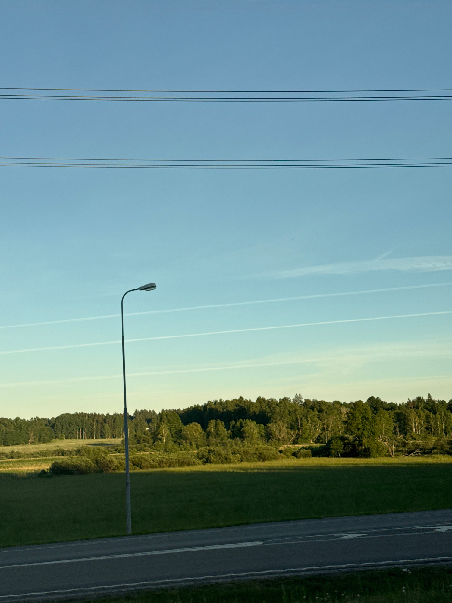 A wide-angle shot captures a serene rural landscape under a clear blue sky. In the foreground, a paved road with white lane markings runs horizontally across the bottom of the frame. To the left of the road stands a tall, slender lamppost. Beyond the road, a lush green field stretches out, leading to a dense forest that forms a tree line across the middle ground. Faint white contrails streak across the sky, suggesting the passage of aircraft. The overall impression is one of tranquility and natural beauty.