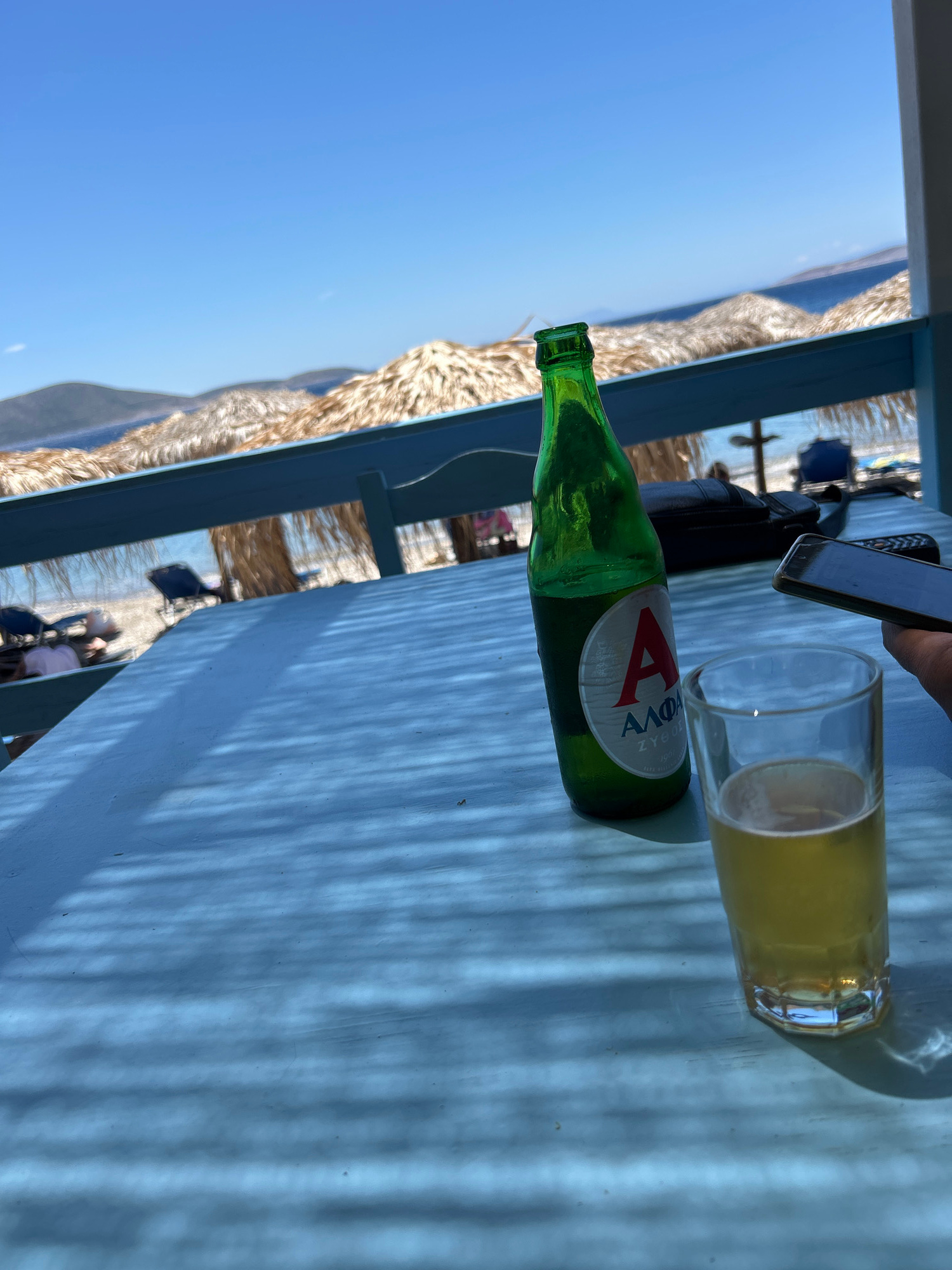 The image captures a vibrant scene at a beachside establishment, likely a bar or cafe. In the foreground, a light blue table is set with a green glass bottle of Alfa beer and a nearly full glass of beer. The bottle has a white label with a red 'A' and Greek text. A dark object, possibly a bag, rests on the table behind the bottle, and a smartphone is held in a hand to the right, suggesting someone is actively using it.
The background reveals a picturesque beach with thatched umbrellas providing shade. Beyond the umbrellas, lounge chairs are visible on the sand, and the tranquil blue sea stretches towards distant hills under a clear, bright sky. The overall ambiance is relaxed and summery, evoking a vacation or leisure setting. The lighting suggests it's daytime, with dappled shadows cast on the table from the overhead structure.