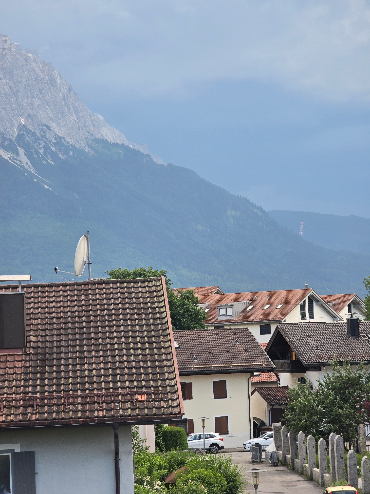 This is a tilted, landscape photograph taken from a high vantage point, likely a balcony or window, looking down and across a residential area nestled against a mountain backdrop. The scene depicts a European village with terracotta-tiled roofs and cream-colored buildings. In the foreground, the roof of a building dominates the right side of the frame, covered in weathered, dark terracotta tiles. A satellite dish is mounted on this roof, facing left.
Further back, a row of houses with similar tiled roofs are visible, their facades featuring windows and dormers. The architecture suggests a European setting, and the context indicates Garmisch-Partenkirchen, Germany. A line of weathered stone pillars, possibly part of a fence or a decorative element, runs along the right edge of the image.
Two white cars are parked on a street or driveway in the middle ground. Lush green trees and bushes are interspersed among the buildings, indicating a summer or late spring setting. In the distance, a massive, imposing mountain range rises, its slopes covered in trees on the lower parts and rocky, bare rock at higher elevations. The sky is a pale, hazy blue, suggesting a slightly overcast or hazy day, or possibly the effect of atmospheric perspective looking towards distant mountains.
High on the mountainside in the far distance, a structure resembling a cable car station or a series of gondolas is faintly visible against the muted blue of the mountain. The overall impression is one of a peaceful, scenic mountain town, with a blend of residential architecture and natural beauty. The time of day appears to be daytime, with soft, diffused light. There is no discernible text in the image.