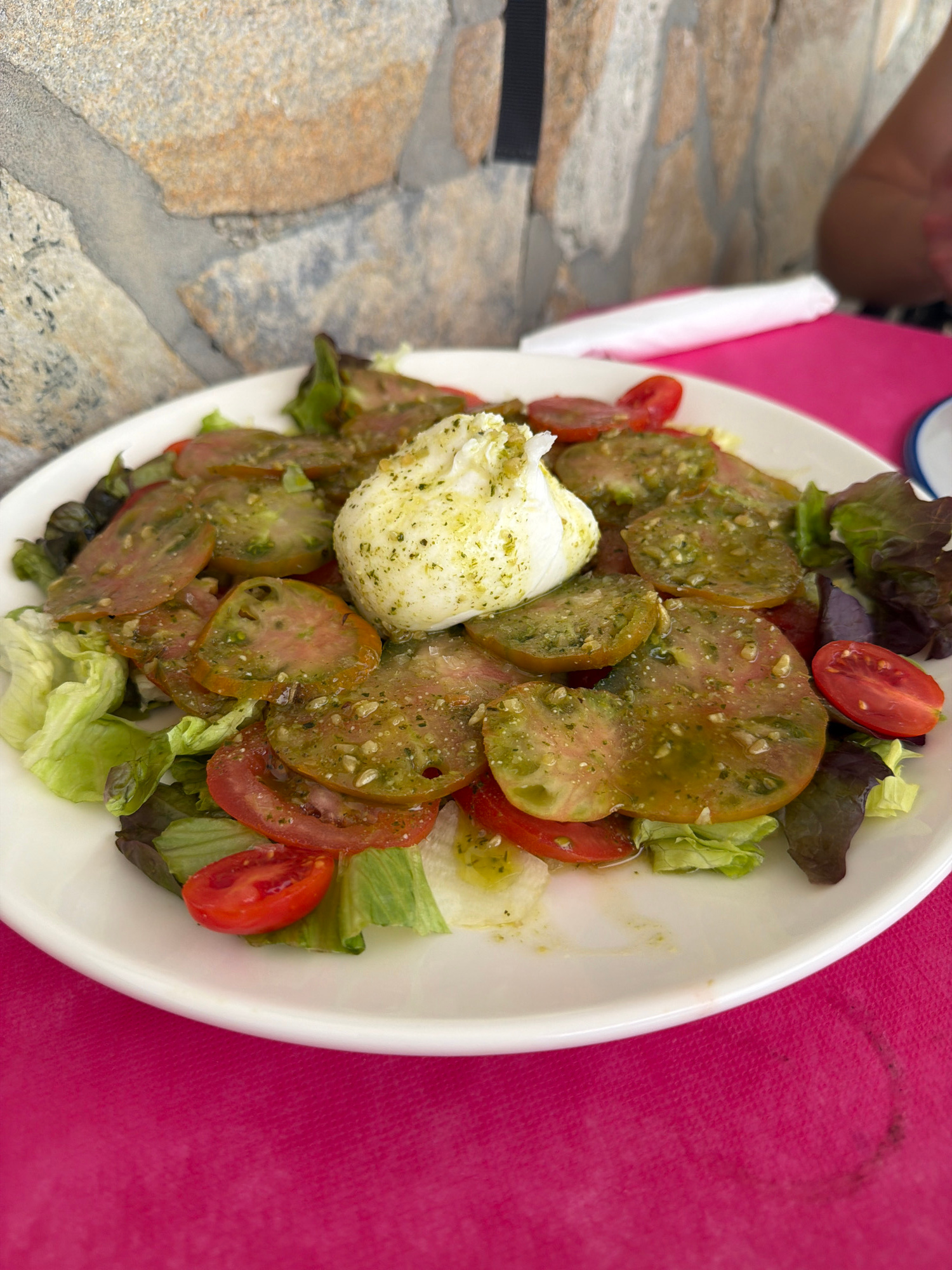 A white plate of Caprese salad is presented on a vibrant pink tablecloth. The salad features sliced heirloom tomatoes, some with a green and pink hue and others bright red, interspersed with crisp green lettuce leaves. A large ball of fresh mozzarella cheese sits prominently in the center of the salad, drizzled with a green herb dressing that also coats the tomatoes. The salad is on a stone-tiled surface, suggesting an outdoor setting. The lighting appears to be natural daylight, indicative of a daytime meal. The background is slightly blurred, focusing attention on the food. The scene evokes a sense of a leisurely meal in a Mediterranean location, consistent with the provided location context of Cabo De Gata, Spain. No visible text is present in the image.