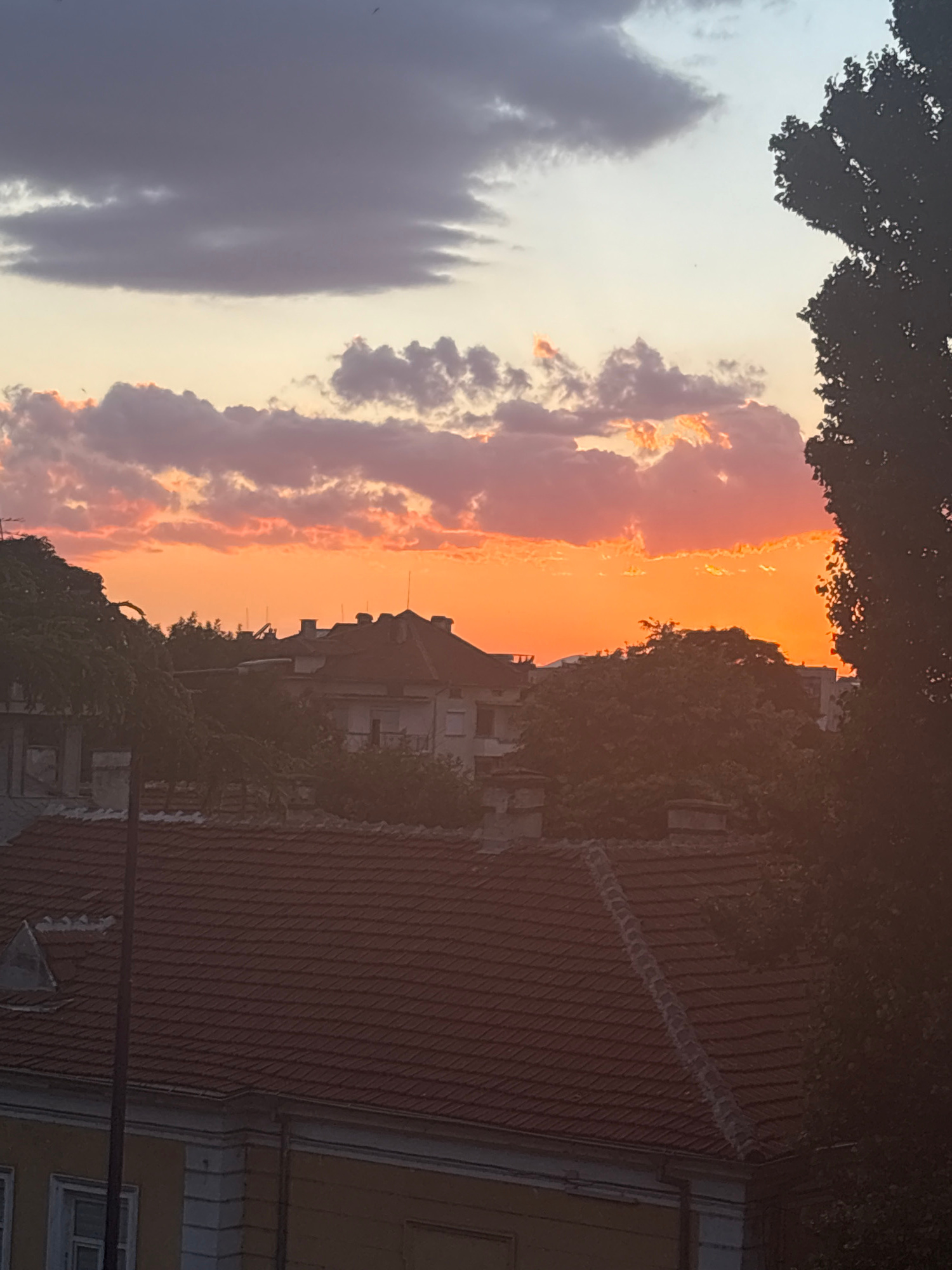 This is an image taken at sunset in Pazardzhik, Bulgaria. The sky is filled with dramatic clouds, illuminated by the setting sun in hues of orange and pink, with darker gray clouds dominating the upper portion of the sky. Silhouetted against the colorful sky are the rooftops of buildings and the dense foliage of trees. In the foreground, a tiled roof and part of a yellow building are visible. The overall scene depicts a tranquil urban landscape at dusk.