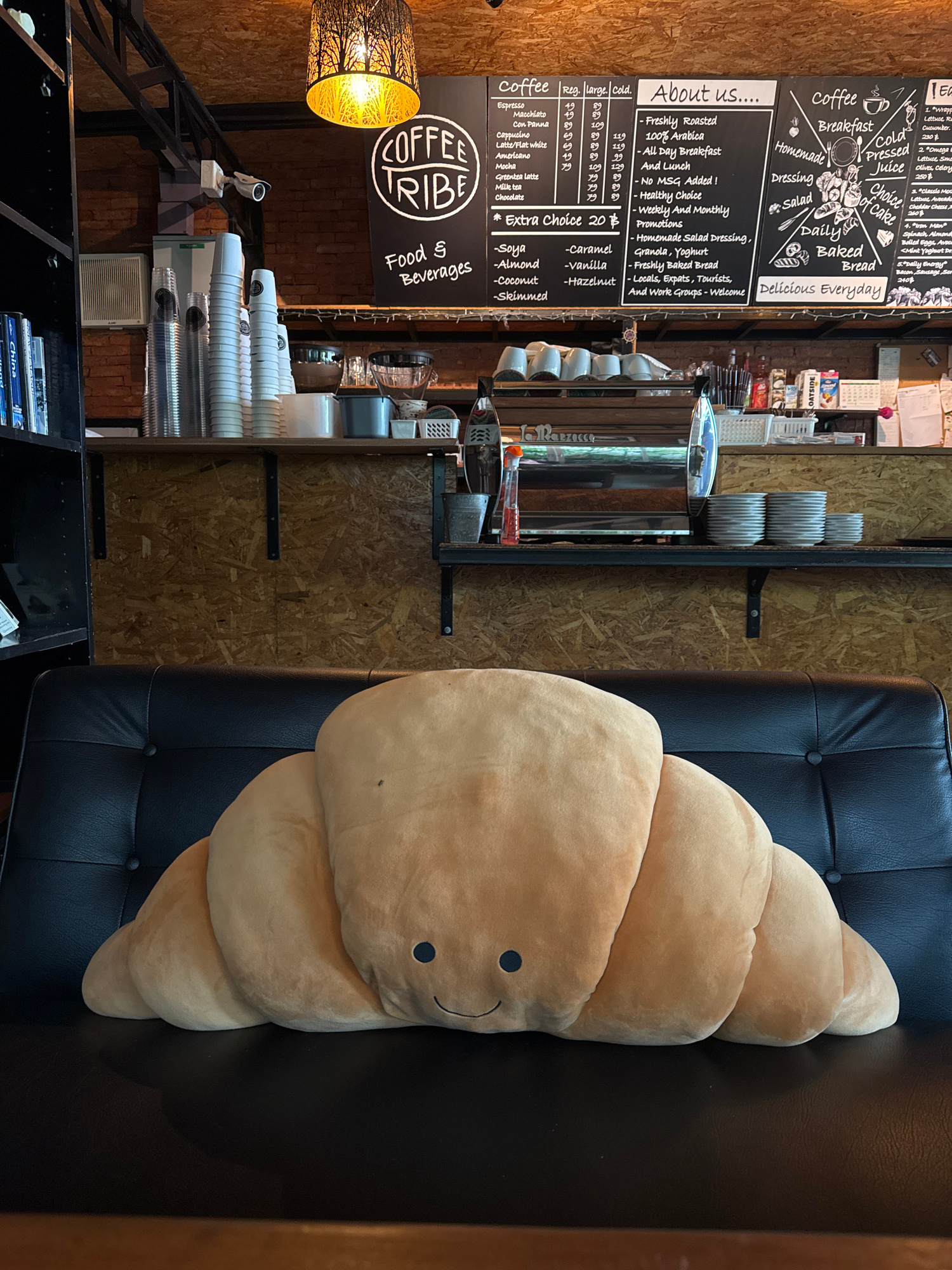 The image shows a cozy cafe interior in Rawai, Thailand, with a large, plush croissant-shaped pillow resting on a black leather sofa in the foreground. Behind the sofa, a shelf displays stacked white cups and a coffee machine. Above the shelf, a blackboard menu lists coffee drinks, food options, and cafe information. A circular logo with the text "COFFEE TRIBE" is visible on the blackboard. The cafe has a rustic ambiance with exposed brick walls and wooden panels.