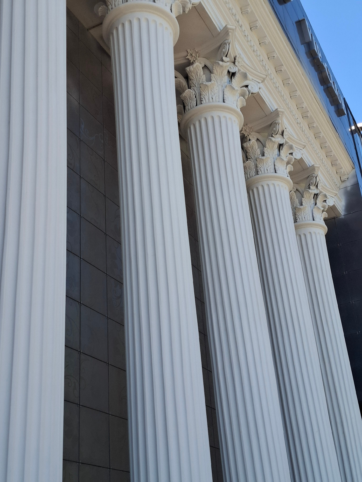 The image displays a close-up, low-angle view of architectural elements, specifically fluted columns and decorative moldings. The columns are white and appear to be made of a smooth material, possibly stone or a composite. They are positioned in a row, extending diagonally across the frame from the bottom left to the top right. The fluting, or vertical grooves, on the columns is clearly visible and adds a sense of texture.
Above the columns, there is an area with intricate white decorative carvings, likely part of the building's facade. These carvings are detailed with floral or acanthus leaf motifs. Further up, a dark, reflective surface is visible, which could be a modern building with dark glass or metal panels. This reflective surface shows a blurred reflection of some white shapes, possibly letters or architectural features. The sky, a clear light blue, is visible in the upper left corner, suggesting it is daytime.
The lighting suggests a sunny day, with light casting subtle shadows and highlighting the textures of the columns and carvings. The scene appears to be an exterior architectural detail of a grand building, possibly a public institution, a museum, or a historic structure. There is no discernible activity or event taking place; the focus is solely on the static architectural features. No people are visible in the image. The location context is Donetsk, Ukraine, but this information is not visually discernible from the image itself. No text is clearly visible or legible.