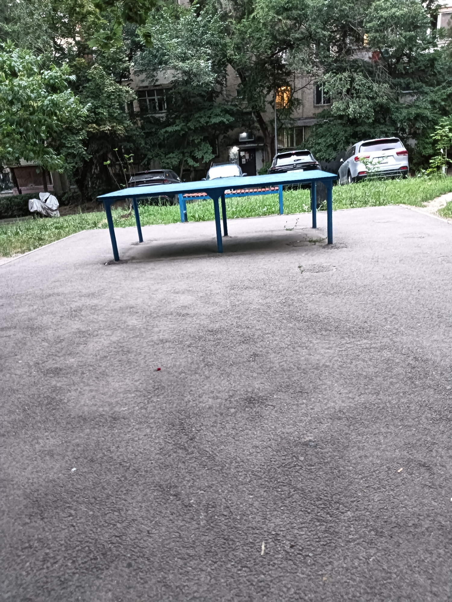 A blue table, possibly a ping pong table, stands on an asphalt surface. Behind it, a building with several windows is partially obscured by lush green trees. Several cars are parked in front of the building. The scene appears to be an outdoor area in a residential complex in Almaty, Kazakhstan.
