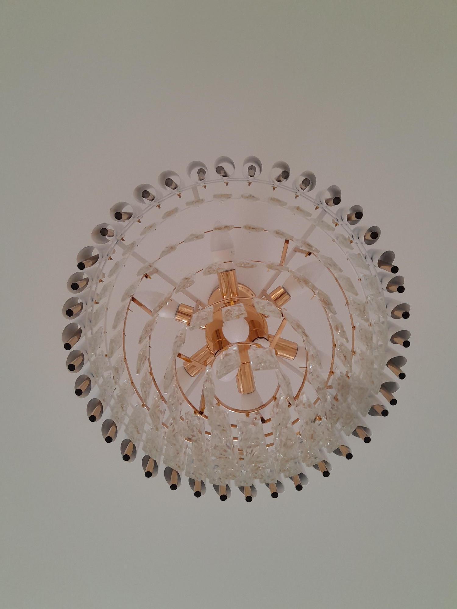A low-angle, full shot captures a modern, circular chandelier against a plain white ceiling. The chandelier features multiple tiers of clear, faceted glass accents arranged in a decorative, leaf-like pattern. These glass elements hang from thin, gold-toned metal frames. The outer edge of the chandelier is lined with small, conical shades, each housing a visible light bulb with a dark tip. The central structure of the chandelier also appears to be made of polished gold-toned metal, with visible light bulbs. The lighting is soft and even, suggesting ambient room lighting rather than direct illumination from the chandelier itself. The overall impression is one of elegant, contemporary design.