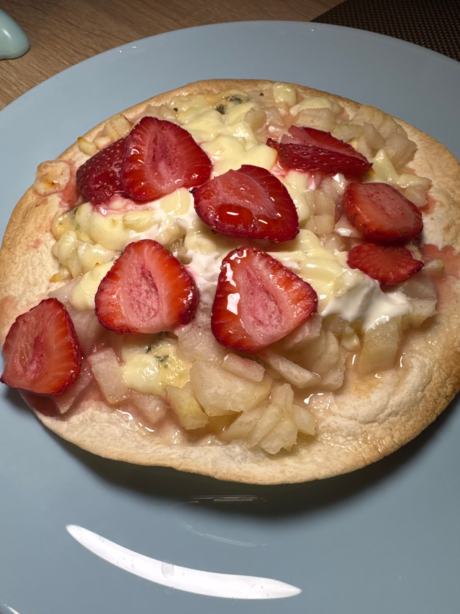 A close-up, high-angle shot captures a small, round flatbread pizza topped with sliced strawberries, pieces of what appears to be pear or apple, and a creamy white sauce, possibly cheese or a béchamel. The pizza has a golden-brown crust and is presented on a light blue plate. The background shows a portion of a wooden surface and a patterned placemat. The location is not specified within the image.