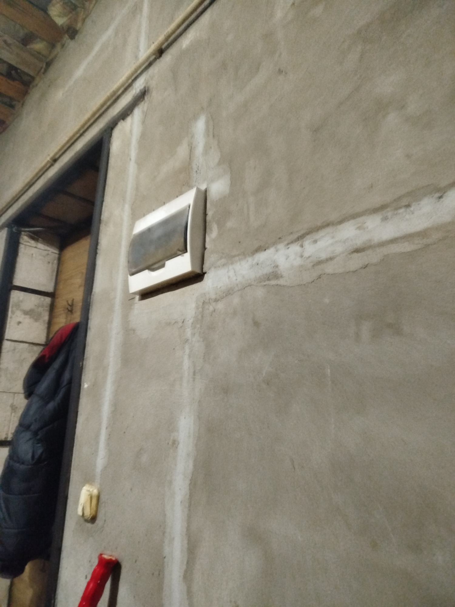 A close-up, angled shot shows a white electrical box mounted on a rough, gray, concrete wall. The box has a clear, hinged cover that is slightly fogged. A metal conduit runs horizontally above the box, and a pipe follows a vertical line down the wall. To the left of the box, a dark blue and red quilted jacket hangs on a dark frame. Below the jacket, a yellow electrical outlet is visible, and a red object with a white tip, possibly a tool handle, rests against the wall. The ceiling above is visible with wooden beams. The scene appears to be an indoor, unfinished space.