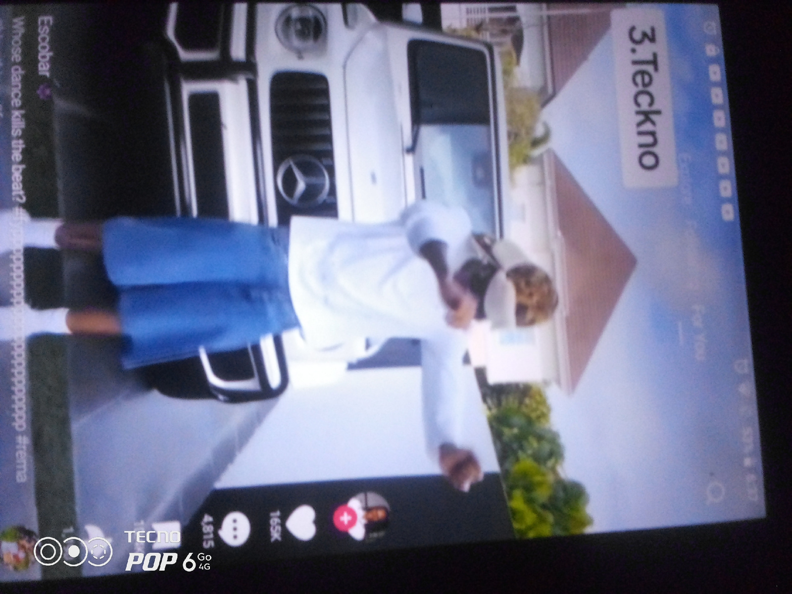 The image is a screen capture from a mobile device, likely a TikTok video, viewed at an angle. The content displayed features a person in white clothing and blue shorts dancing in front of a white vehicle that resembles a Mercedes-Benz G-Wagon. Text overlays are present, indicating a ranking of "3. Teckno" and a username "Escobar" with the accompanying text "Whose dance kills the beat? #rema". The bottom of the screen shows interface elements typical of a social media application, including icons for likes, comments, shares, and user profiles, along with branding for "TECNO POP 6 Go 4G". The background suggests an outdoor setting with greenery and a building. There is no specific indication of the location being Howayi, Nigeria.