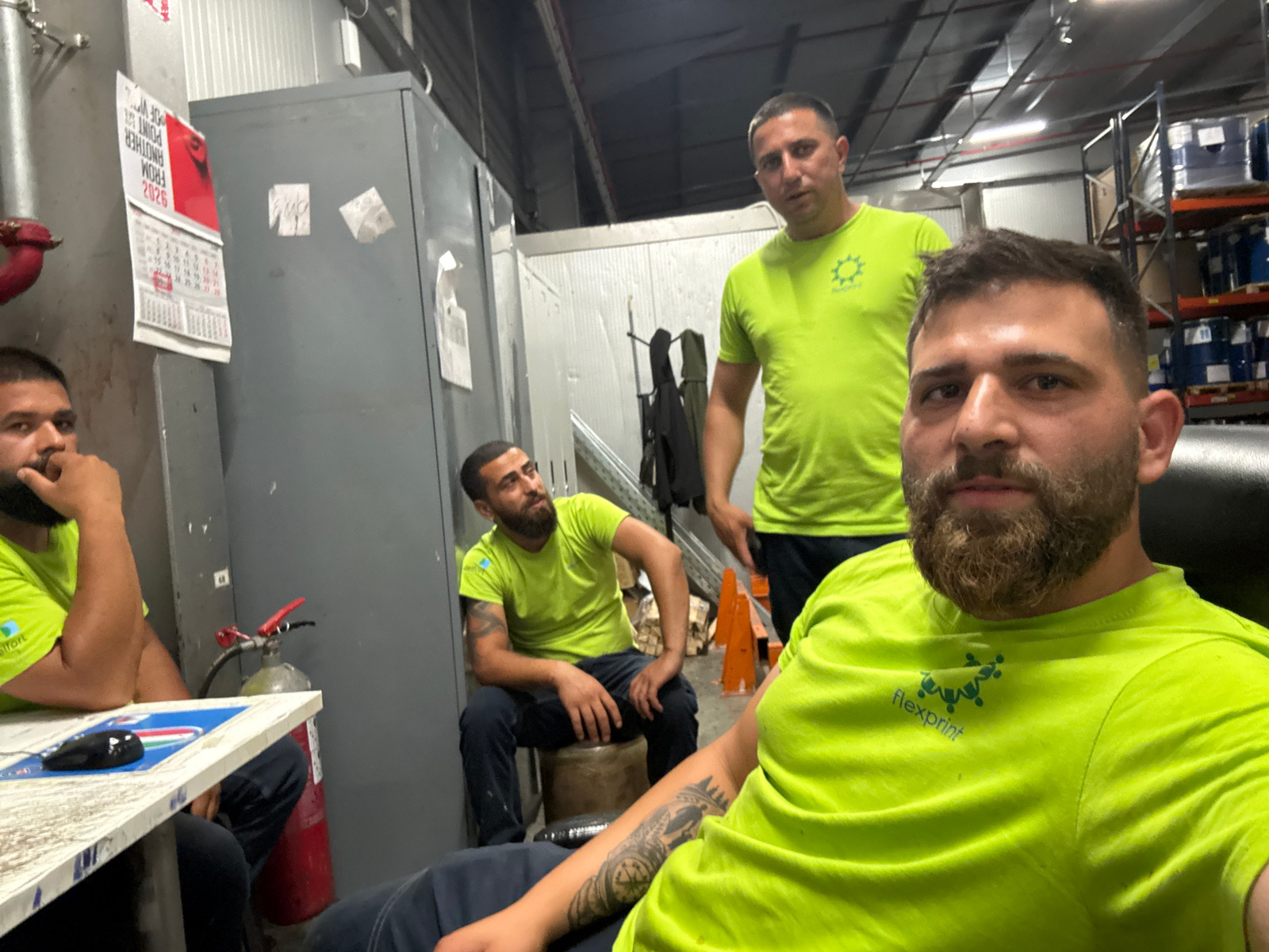 Four men are in an indoor industrial setting. Two men in the foreground are wearing bright green t-shirts with a logo that reads "flexprint." One is looking directly at the camera, while the other has his hand on his chin. To the left, a third man is sitting at a table and looking away from the camera. He is also wearing a bright green t-shirt. In the background, a fourth man in a similar t-shirt is standing and looking towards the camera.
The setting appears to be a warehouse or workshop, with industrial shelving units stacked with blue barrels visible in the background. A large gray cabinet is on the left, with a red fire extinguisher nearby. A calendar is hanging on the cabinet, showing the month of May and the year 2026. The lighting suggests it is either late in the day or indoors with artificial lighting. The overall mood seems casual, with the men possibly on a break.