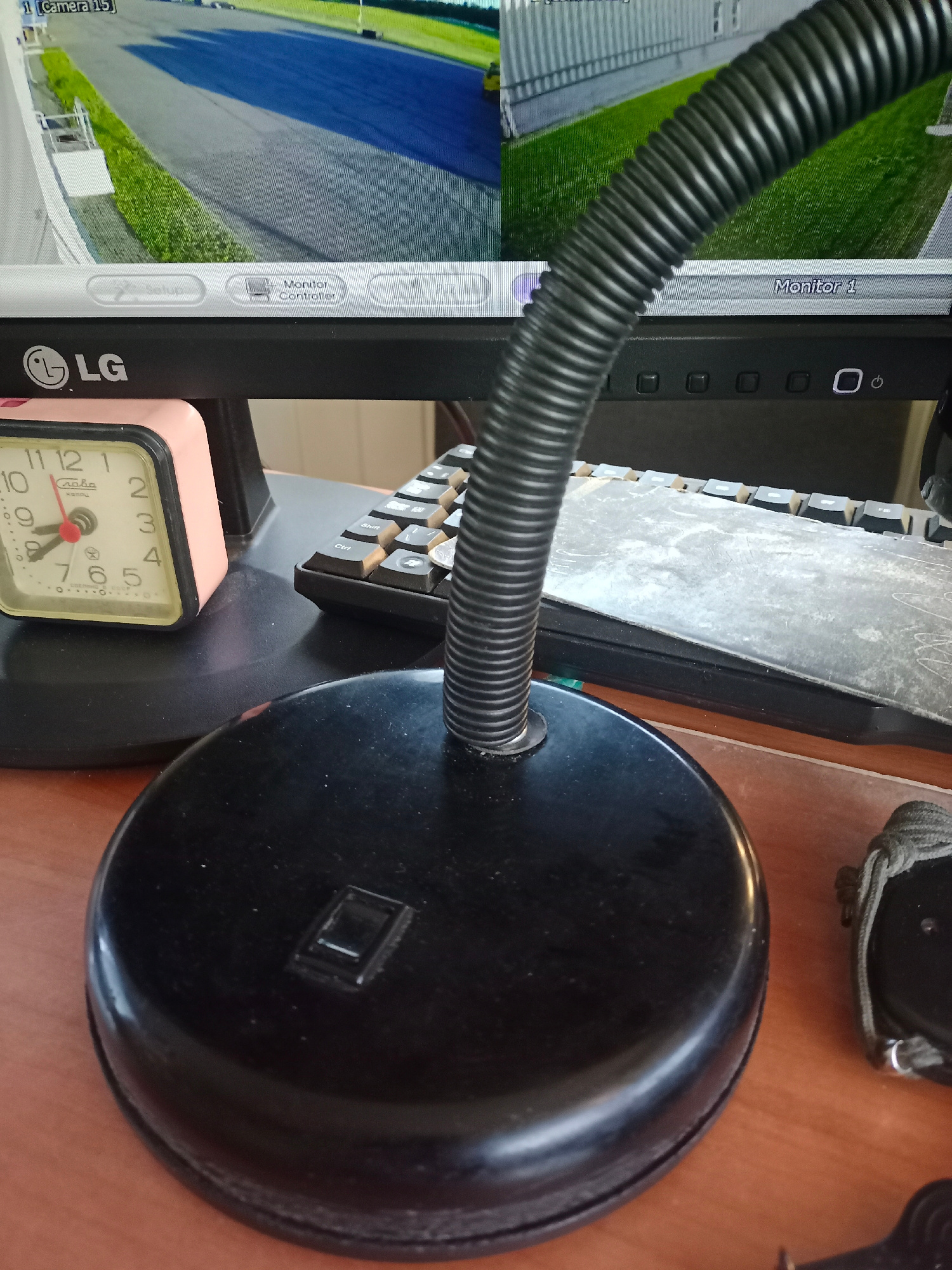 The image shows a close-up of a desk with various objects, including a computer monitor displaying what appears to be security camera footage of an outdoor area with a race track and grass. Below the monitor, an LG logo is visible. To the left of the monitor, a pink analog alarm clock is placed on a black circular base. A black gooseneck lamp extends from another black circular base in the foreground. Part of a keyboard is visible to the right of the clock, with a protective cover over some of the keys. To the far right, a black object with a braided strap is partially visible. The scene is set on a wooden desk surface. The location is identified as Shchomyslitsky Sel'sovet, Belarus. There are no people present in the image, and no actions or interactions are depicted.