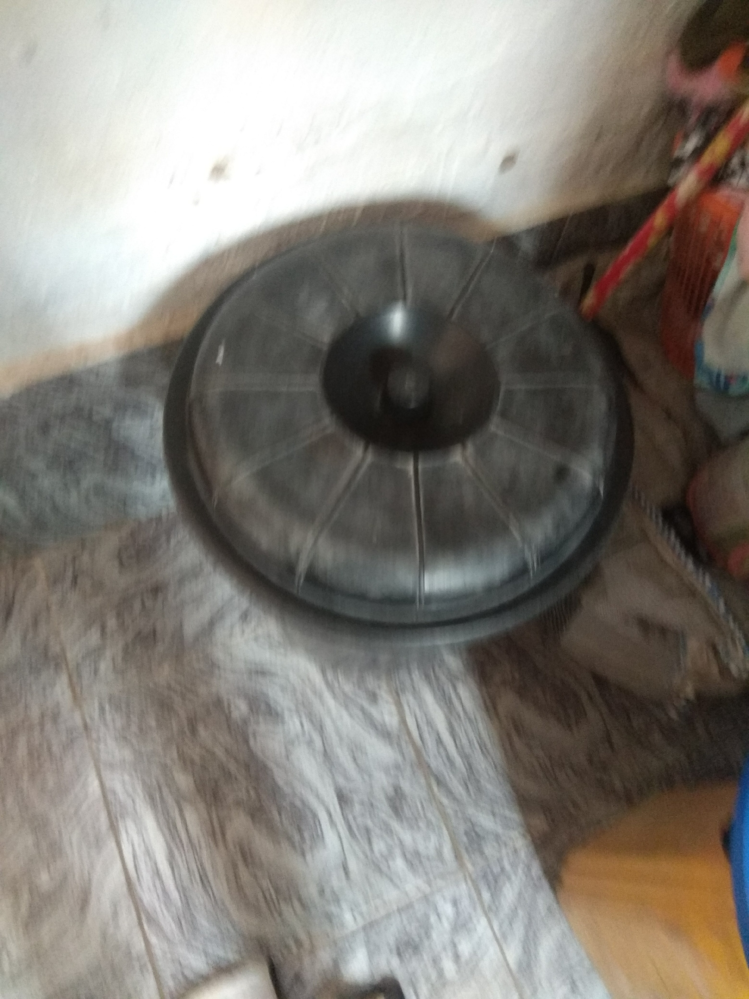 The image shows a black, circular object with a textured surface, possibly a lid or a shallow basin, resting on a patterned floor. The object has radiating lines emanating from a central darker circular area. To the right of the object, partially visible, are colorful items that appear to be a broom with red and yellow stripes and some fabric or bags. The background features a light-colored wall with some darker marks. The floor has a tiled or linoleum pattern with shades of gray and white, and a roughly textured brown area is visible to the right of the black object. The lighting suggests an indoor setting, possibly dimly lit, with shadows cast by the objects. No people are visible. The scene appears to be an arrangement of household items in a room, with no specific activity depicted.