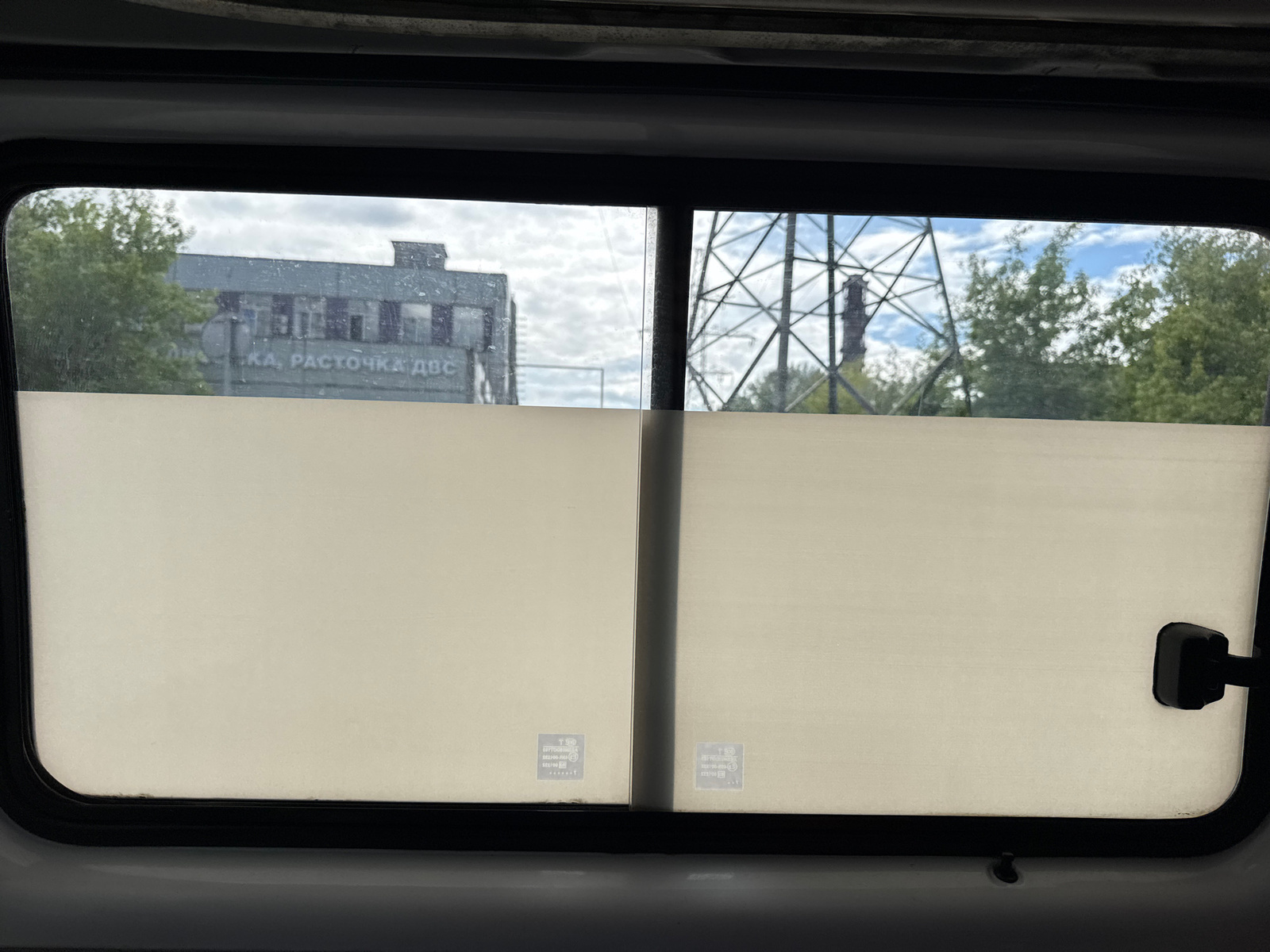 The image shows a view from inside a vehicle looking out a window. The window is divided by a vertical bar, with two frosted or translucent sunshades covering the lower portion of the window. Through the upper, clear portion of the window, a weathered industrial building with the Cyrillic text "КА, РАСТОЧКА ДВС" (which translates to "Engine Honing") is visible to the left. To the right, a large metal lattice tower and a smokestack are present, along with trees and a cloudy sky. There is a handle on the right side of the window frame. The scene appears to be in an industrial or commercial area.