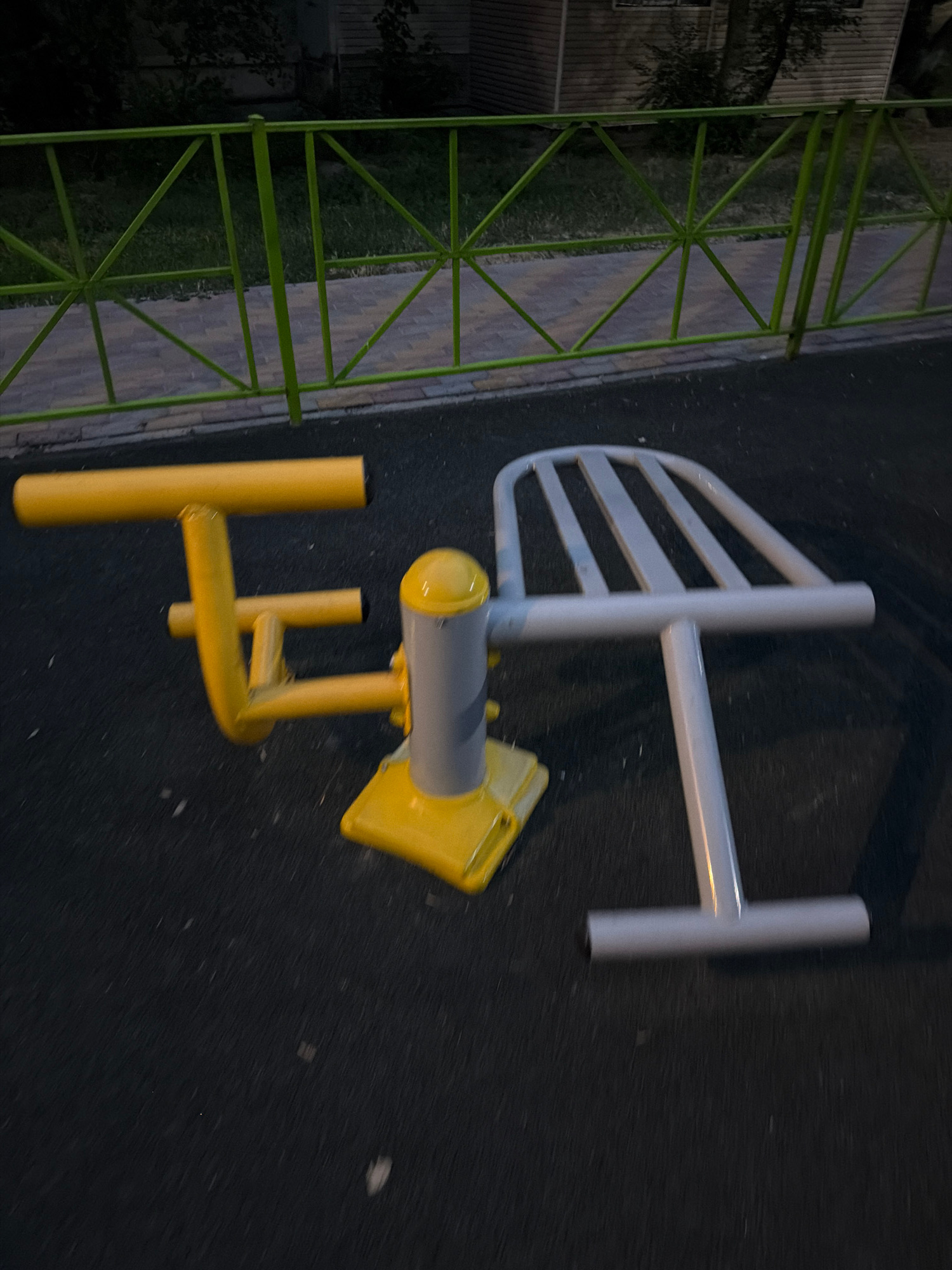 This media file captures an outdoor exercise machine on a paved surface, likely a park or public fitness area, in Shymkent, Kazakhstan. The scene appears to be set at dusk or night, as indicated by the low light conditions and the dark, reflective asphalt. The exercise equipment is primarily yellow and white, with some green elements visible from a nearby fence or railing.
The main subject is a piece of outdoor gym equipment designed for abdominal exercises, featuring a curved backrest and handgrips. A yellow, seat-like component is attached, suggesting it's a sit-up bench or a similar apparatus. The machine is made of metal, with a robust construction suitable for outdoor use. A green metal fence, with a geometric lattice design, borders the area on the left side of the frame. The fence is likely part of the park's infrastructure. The ground is a dark, asphalt surface, speckled with small white debris, possibly salt or dirt.
There are no people visible in the frame, and the focus is solely on the equipment and its immediate surroundings. The time of day is late, as indicated by the artificial light casting shadows and illuminating parts of the equipment. The weather appears to be clear, with no visible rain or snow.
The overall impression is one of an accessible public space designed for fitness. The location cues, such as the specific type of outdoor gym equipment and the style of fencing, are consistent with public recreational areas. No text is visible in the image.