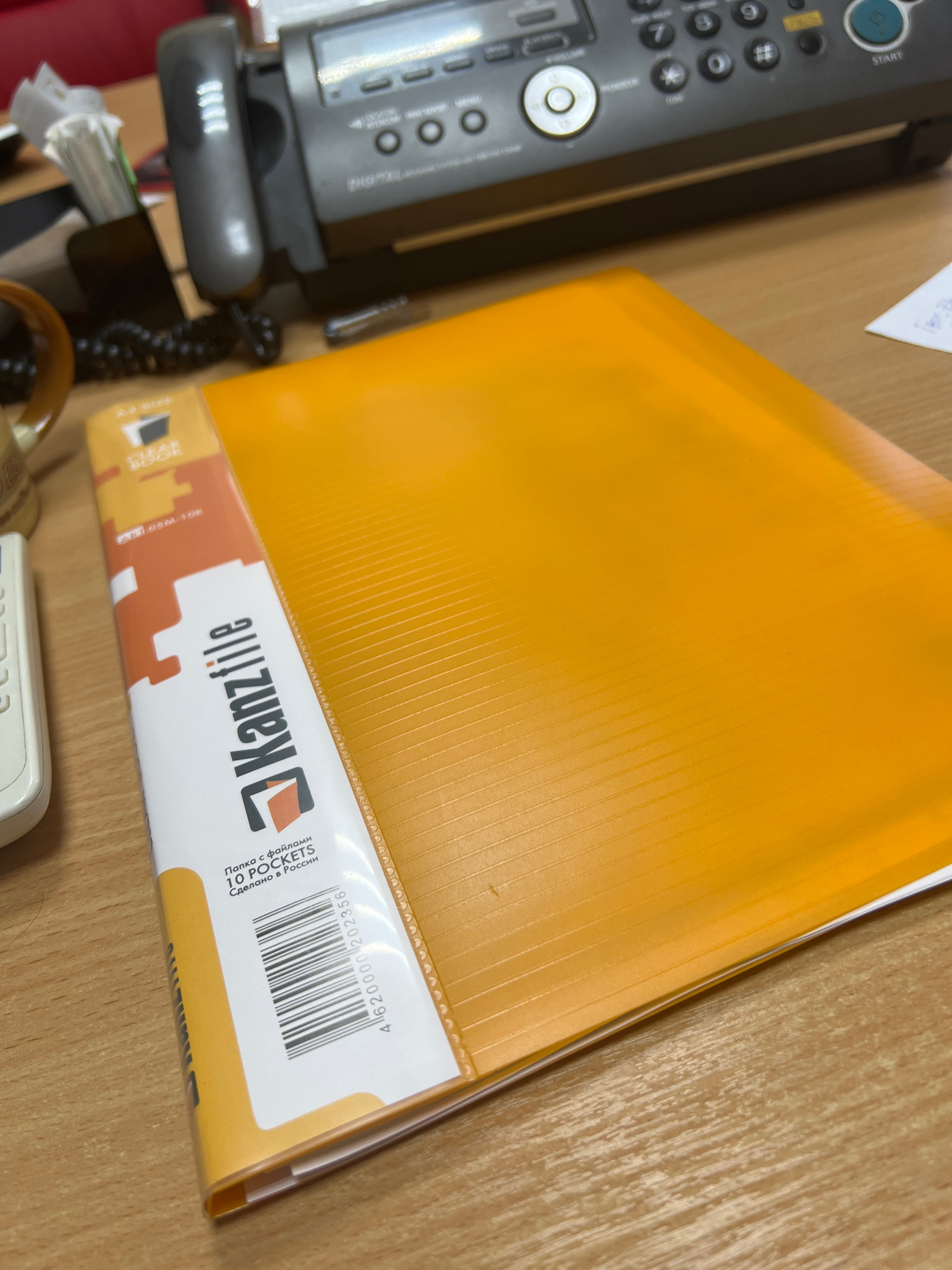 An orange plastic binder with a white label featuring the brand name "Kanzfile" and "10 POCKETS" is placed on a wooden desk next to a fax machine. The binder is open at the bottom, showing the plastic pockets. A barcode and Russian text are visible on the label. The setting appears to be an office or home office environment. There is no specific information about the location in Tula, Russia.