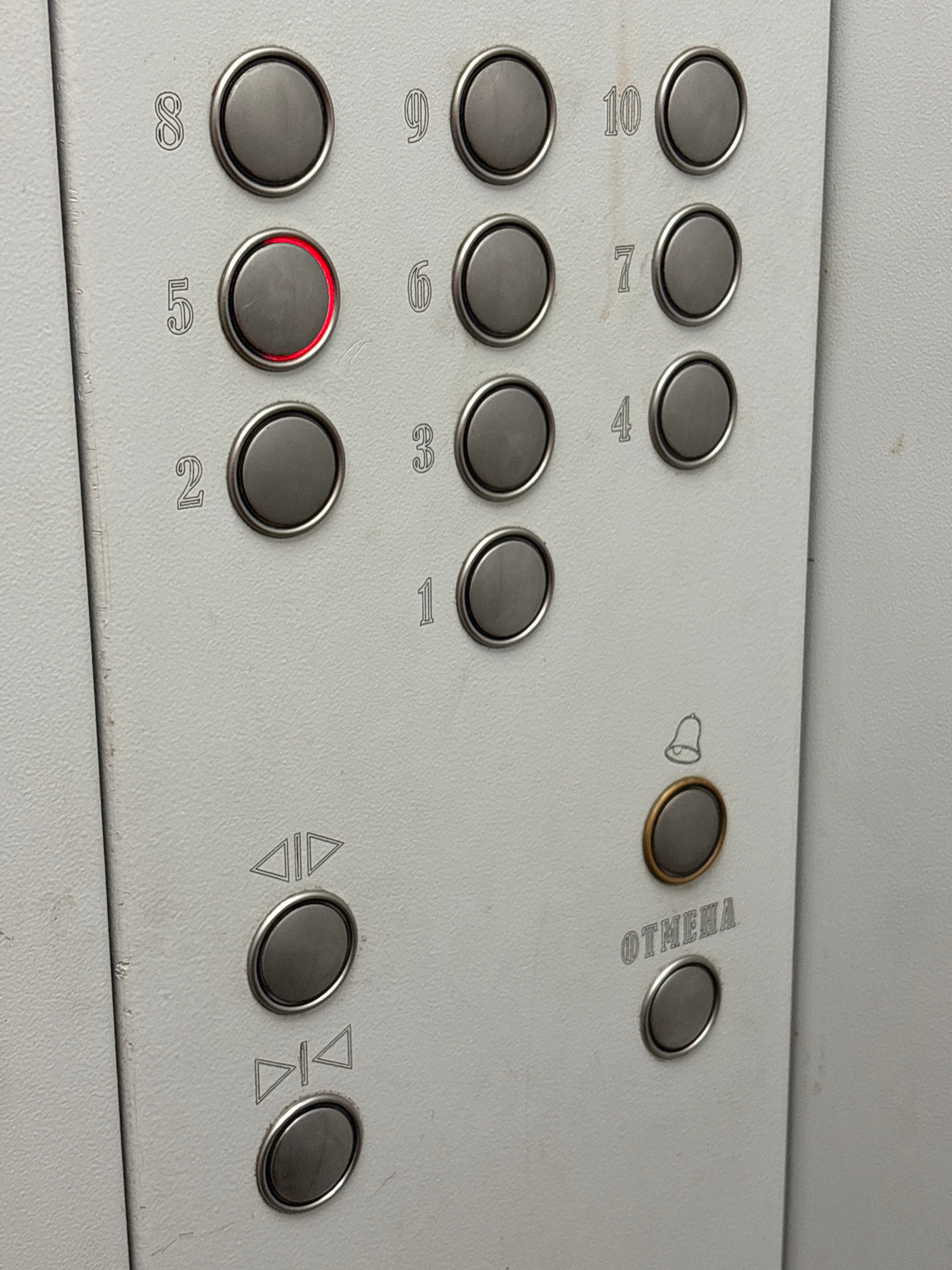 The image shows a close-up view of an elevator control panel. The panel is made of a light-colored material, possibly metal, and is marked with buttons and symbols. Several numbered buttons are visible, ranging from 1 to 10, arranged in two columns. The button labeled "5" is illuminated with a red light. Below the numbered buttons, there are two buttons with arrow symbols, indicating "up" and "down." Further down, there is a button with a bell symbol and another button with the Russian word "ОТМЕНА" (OTMENA), which translates to "cancel" or "recall." The buttons are round and appear to be made of metal. The overall condition of the panel suggests it is in use, with some minor scuffs or marks on the surface. The background is a plain, light-colored wall, likely the interior of an elevator shaft or hallway. There are no people visible in the image. The presence of Cyrillic script and the style of the elevator panel might suggest a location in Belarus, but this specific location in Mahilyow cannot be determined from the image alone.