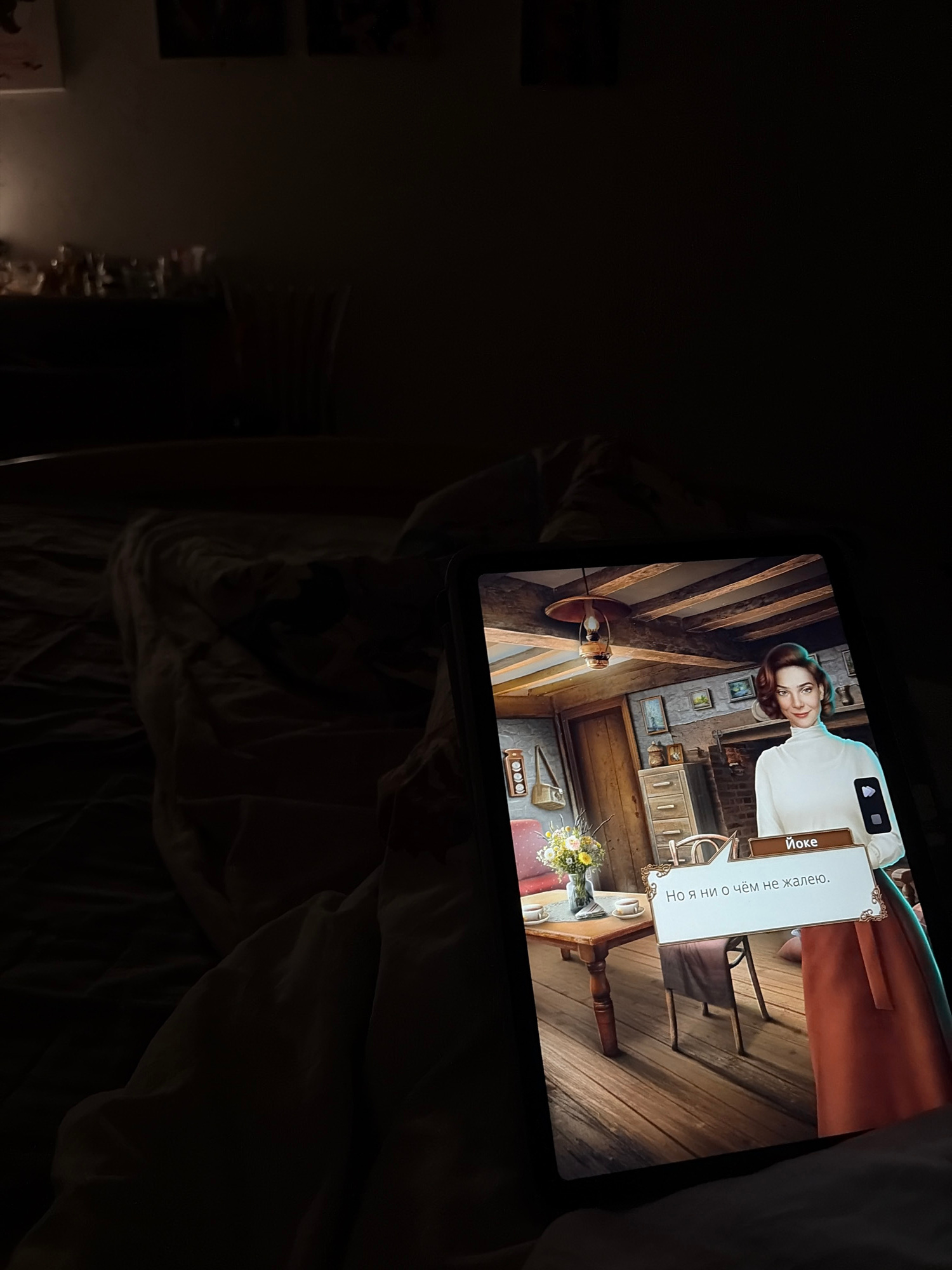 An image captures a tablet screen displaying a scene from a video game. The game features a woman in a white turtleneck and an orange skirt, set in an interior that resembles a rustic room with wooden floors and ceiling beams. A speech bubble on the screen contains text in Cyrillic, and the name "Йоке" is visible. The tablet is held by an unseen person, and the background is dimly lit, suggesting an indoor setting, possibly a bedroom. The image's context does not provide any information that would allow for identification of a specific location within the city of Dnipro, Ukraine.