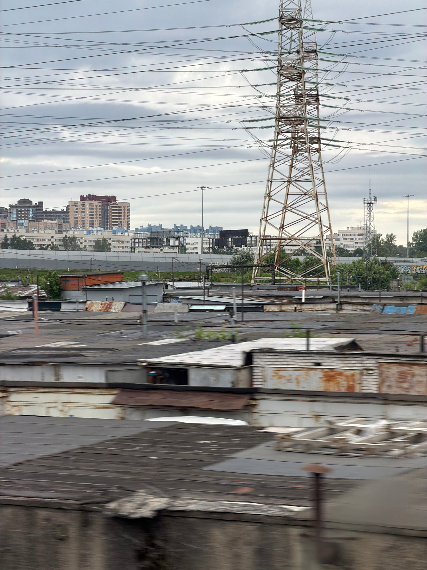 The image captures an urban landscape under a cloudy sky, dominated by a tall, lattice-style electricity pylon. Numerous power lines crisscross the frame, creating a dense network against the muted sky. In the foreground, a series of low-rise, weathered structures, likely garages or sheds with corrugated metal roofs, are visible. Some of these structures show signs of rust and age. Beyond these, a wider cityscape unfolds with a cluster of multi-story apartment buildings in varying shades of beige, brown, and white. Some buildings have visible windows and architectural details. In the middle ground, a chain-link fence or barrier runs horizontally, separating the foreground structures from the background buildings. Another, smaller utility tower is visible in the distance to the right of the main pylon. The overall impression is one of an industrial or semi-industrial area on the outskirts of a city, with evidence of aging infrastructure and residential development. The motion blur suggests the image was taken from a moving vehicle.