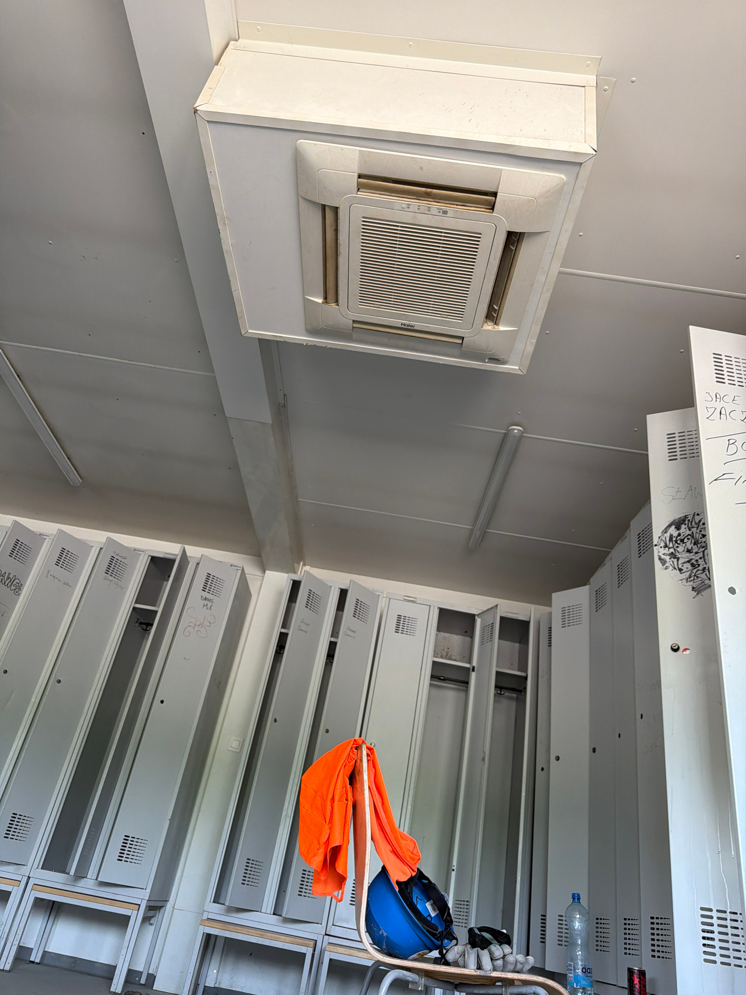 The image shows an indoor scene, likely a storage area or locker room, with a focus on a ceiling-mounted air conditioning unit on the left and rows of metal lockers on the right. The air conditioning unit is white and rectangular with a grille in the center. Above it, a section of a white panel is visible, with some handwritten text and markings on it.
The lockers on the right are arranged in vertical stacks, with some doors open and others closed. They are made of light-colored metal and have ventilation grilles on the front. On one of the lockers, an orange piece of fabric, possibly clothing, and a blue hard hat are placed on top, suggesting recent activity or storage of safety equipment. A water bottle is also visible on a shelf within the locker unit.
The overall impression is one of a functional, utilitarian space. The lighting appears to be artificial, casting a neutral light on the environment. The image is taken from a slightly elevated angle, looking down and across the room. There are no people visible in the image. The visible text includes "Haier" on the air conditioner, and handwritten names or labels on some of the lockers, such as "JACE," "ZACE," "BO," and "SŁAW." There is also a label that appears to read "IDO."