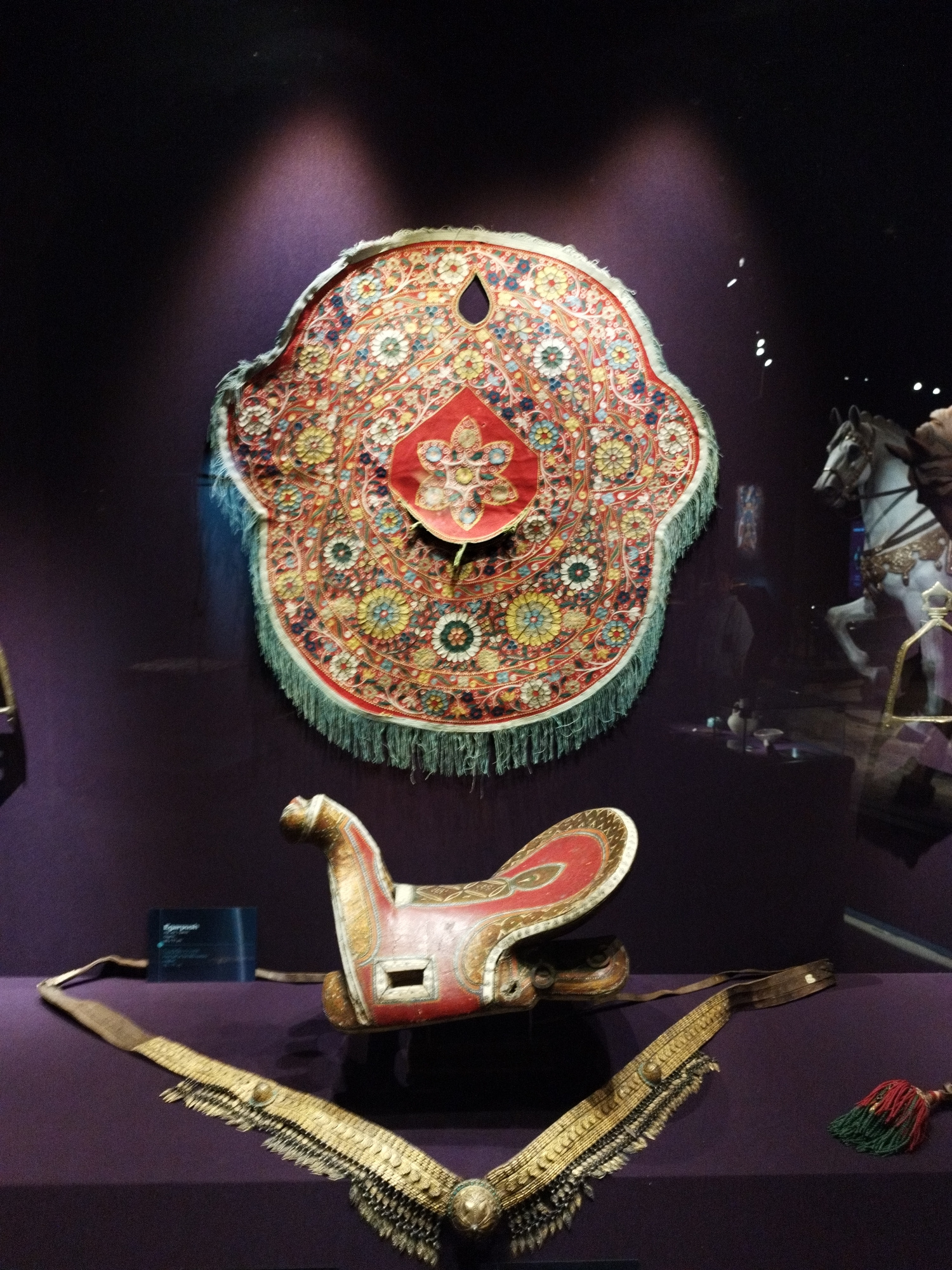 The image displays a museum exhibit featuring equestrian artifacts, likely from a historical or cultural display. In the foreground, a red and gold saddle is positioned centrally, with a decorative gold and black strap laid out in front of it. Behind the saddle, a large, ornate fabric tapestry with intricate floral patterns in red, blue, yellow, and white hangs against a dark purple backdrop. The tapestry features a fringe of green threads. To the right of the tapestry, a white model horse, caparisoned and in mid-stride, is visible. The environment appears to be an indoor exhibit with subdued lighting, casting a spotlight on the central tapestry and saddle. The floor and backdrop are a consistent dark purple.
