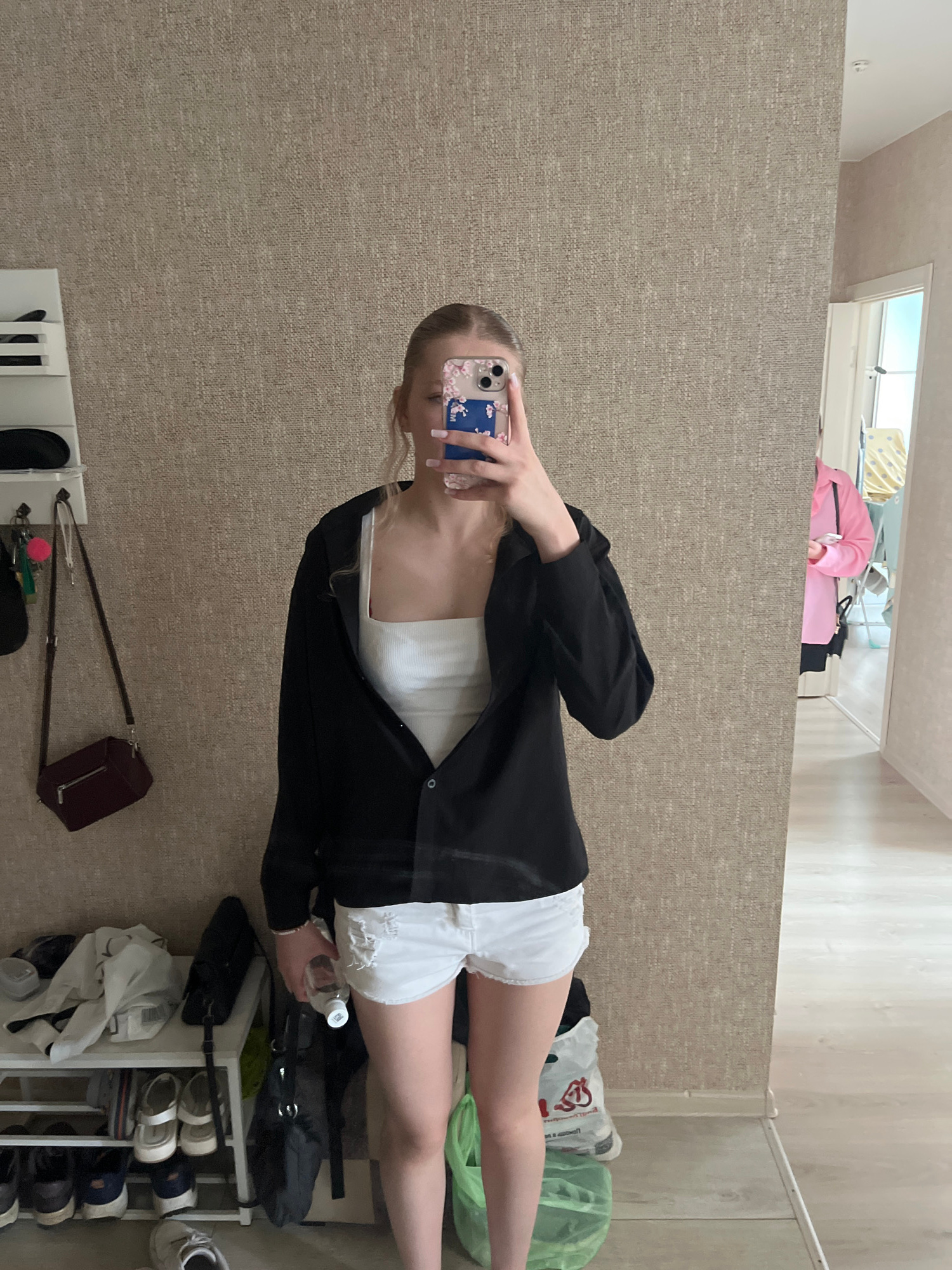 The image shows a young Caucasian woman with blonde hair pulled back in a ponytail, taking a mirror selfie. She is wearing a white tank top, an open black button-down shirt, and distressed white denim shorts. She is holding a clear water bottle in her right hand. Her left hand is holding a smartphone with a floral case.
In the background, to the left, is a white shelving unit with various items, including bags, shoes, and what appears to be a dark red handbag hanging from a hook. To the right of the woman, a doorway leads to another room where another person, wearing a pink shirt and black shorts, is visible. The setting appears to be an indoor space, possibly an apartment or house, with textured wallpaper and a wooden floor. The presence of a shoe rack and items like bags suggests a residential area. The image has a neutral lighting condition. No specific elements in the image conclusively place the scene in Perm, Russia.