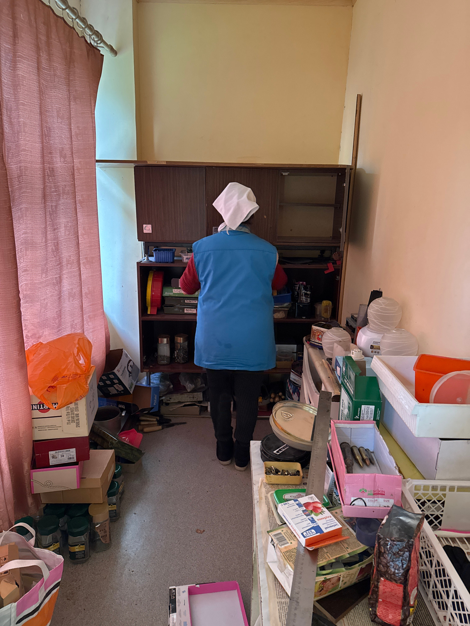 The image shows a cluttered room, possibly a storage area or a workshop, with various items scattered on shelves and the floor. A person wearing a blue jacket and a white cap is visible, facing away from the camera and appearing to be organizing or sorting through items on a shelf. The room has a light-colored wall and a wooden cabinet.
On the shelves, there are many objects, including boxes, containers, and what appears to be food items, such as a bag of coffee beans and a box labeled "Universol Клубни" which likely contains strawberries. There are also tools and electronic devices. The floor is covered with more boxes, plastic bags, and what looks like wine bottles. One prominent text visible is "MARTINI SPARKLING WINE-EXTRA DRY".
The lighting suggests it might be daytime, but there are no windows visible to indicate the weather or specific time of day. The overall impression is one of disarray, suggesting a space that is either in the process of being decluttered or is simply very disorganized. The setting appears to be indoors, likely in a residential or commercial storage space. The presence of numerous items and the act of someone sorting through them imply an ongoing task of organization or cleanup.