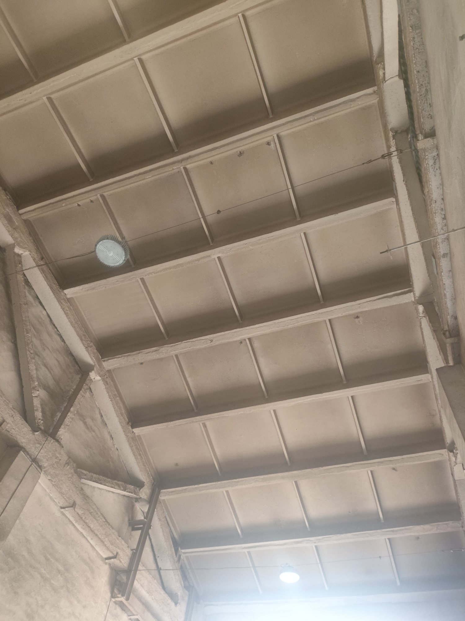The image shows a concrete ceiling structure with visible beams and panels, likely in an industrial or unfinished building. A circular light fixture is suspended from the ceiling by wires, and another light source is visible in the lower right corner, creating a bright glare. The concrete appears weathered and unfinished, with some wires and suspension elements visible. The scene is shot from a low angle, looking upwards at the ceiling. There are no people or discernible objects on the ceiling itself. The environment appears to be indoors, with a utilitarian and unadorned aesthetic.