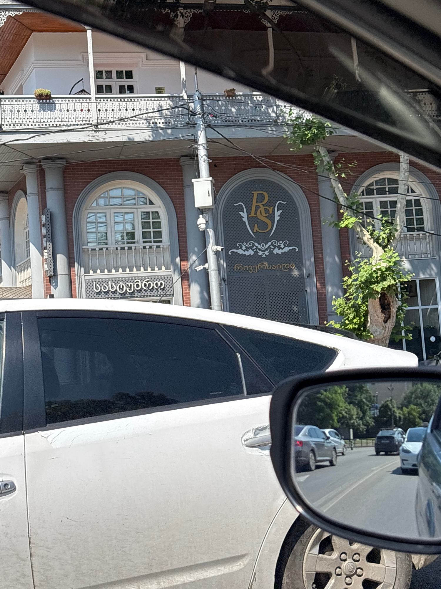 The image is a medium shot capturing a view from a car's interior, looking outward. In the foreground, the side of a white car is visible, with its door handle and a portion of the wheel well. A side-view mirror reflects a street scene with several cars driving on a paved road. Trees and buildings line the road in the reflection.
The background of the image features a multi-story building with a red brick facade and white architectural details. The building has arched windows with intricate grilles and balconies. A prominent logo, "RS," is visible on the facade, along with Georgian script above and below it. The script above the logo reads "სასტუმრო" (gastrosom) and below it reads "ჩიჟინაშვილი" (Chijinashvili). The building appears to be located in the city of Tbilisi, Georgia. The overall scene suggests a sunny day, with shadows cast on the building.