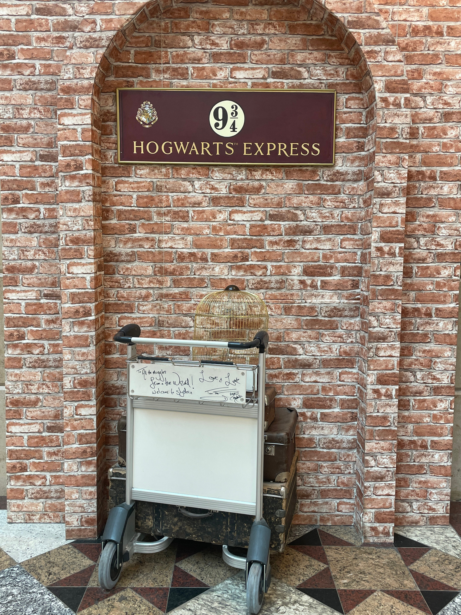 This is a vertical, eye-level, medium shot image of a brick wall with a sign and a luggage trolley. The wall is made of reddish-brown bricks with white grout. To the left, an arched brick alcove frames a rectangular sign with a dark maroon background and a gold border. The sign features a circular emblem with the number "9¾" in gold, and below it, the words "HOGWARTS™ EXPRESS" in gold lettering. The right side of the image shows a luggage trolley with a white board attached. There are several suitcases stacked behind the trolley. A round, gold birdcage sits on a ledge above the trolley. The floor is decorated with a diamond pattern of granite tiles in shades of brown, beige, and black. The lighting suggests an indoor setting, possibly a themed attraction or a replica of a famous location.
Visible text includes "9¾", "HOGWARTS™ EXPRESS", and handwriting on the white board of the trolley that appears to say, "To the Muggles from a the Wizard. Welcome to Slytherin." The handwriting also includes the name "DRACO." The scene evokes a sense of wonder and magic, likely related to the Harry Potter franchise.
