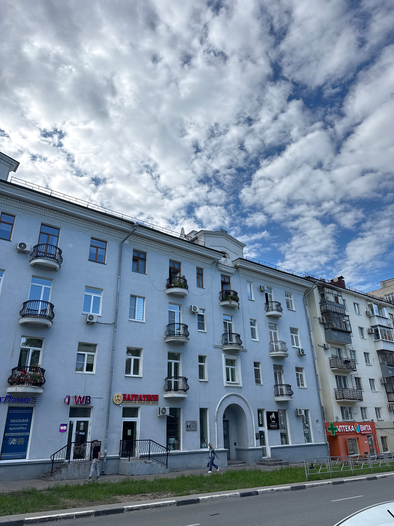 The image captures a street view in Samara, Russia, focusing on the facade of a light blue, multi-story building under a partly cloudy sky. The building features several balconies with ornate black railings, numerous windows, and air conditioning units. Several businesses are visible on the ground floor, including shops with signage such as "WB" and "АПТЕКА ВИТА" (Apteka Vita), an orange pharmacy with a green cross logo. Two individuals are present: one person in a dark shirt and light pants walking on the sidewalk, and another person in a denim jacket and white sneakers walking in front of the pharmacy. The building is situated on a street with a sidewalk and a road in the foreground. The architecture suggests a Soviet-era residential building with commercial spaces at street level.