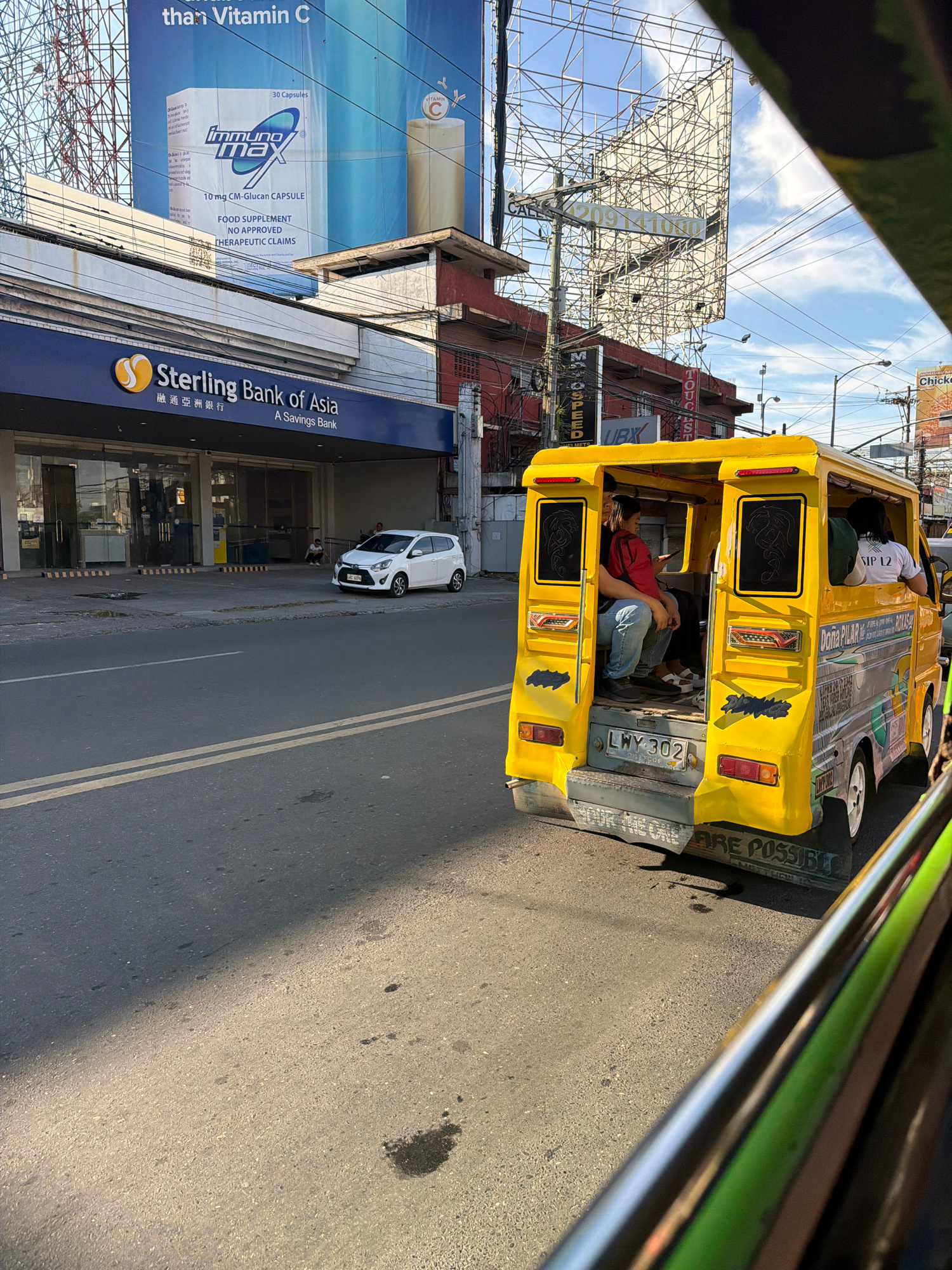 In Davao City, Philippines, a yellow jeepney with passengers is captured mid-motion on a paved road. The jeepney is adorned with colorful decals, including the name "DOÑA PILAR". A white sedan is parked to the left of the frame, in front of a Sterling Bank of Asia building. The scene is illuminated by the sun, casting shadows on the road. The backdrop includes a large billboard advertising "Immuno Max" and various utility poles and wires, indicative of an urban environment.