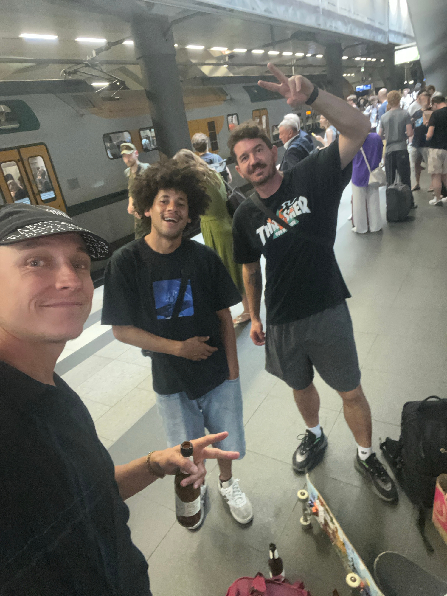 Three individuals are standing on a train platform in Berlin, Germany, with a train visible in the background. The person on the left, wearing a black baseball cap and black shirt, is in the foreground and appears to be taking a selfie. A bottle of beer is visible in his right hand. To his right is another person with curly hair, wearing a black t-shirt and denim shorts, smiling at the camera. The third person, on the far right, is wearing a black t-shirt with a graphic print and grey shorts, and is making a peace sign with his left hand. Other people and luggage are visible on the platform in the background. The platform is illuminated by overhead lighting. A skateboard is lying on the ground near the person on the right.