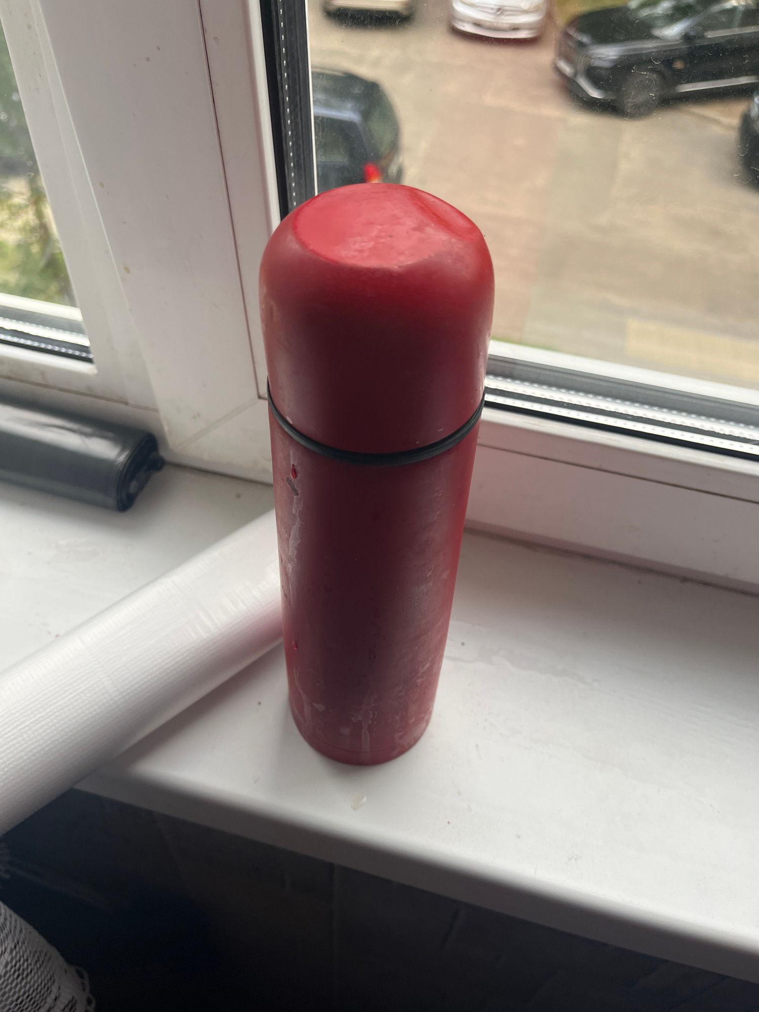 A vertical, slightly angled medium shot shows a red cylindrical object with a black band around its upper half. The object is placed on a white windowsill. To the left of the red object is a roll of white, textured material, possibly plastic wrap, and further to the left, a black, rolled-up object. The windowsill is positioned in front of a window looking out onto a street with parked cars. The background outside the window appears to be a city environment, with buildings and pavement visible. The red object appears to be slightly damp or has condensation on its surface. The overall impression is an indoor scene looking outwards through a window. The objects are arranged in a static composition.
