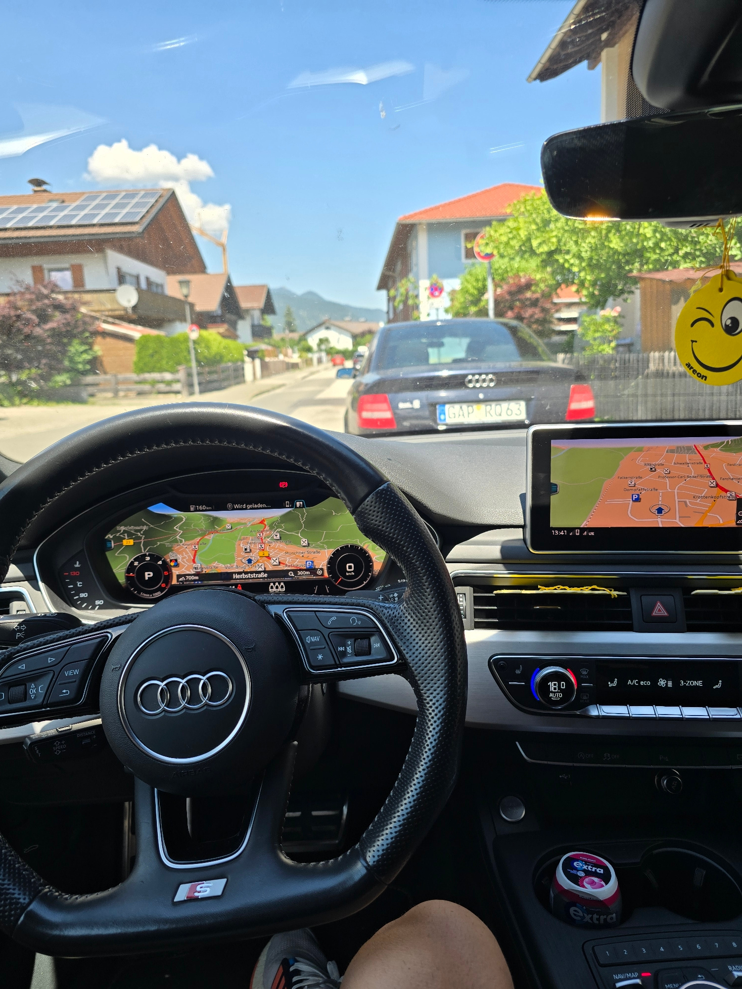 This image is a first-person perspective captured from inside a car. The scene is a residential street in Garmisch-Partenkirchen, Germany, bathed in bright daylight under a clear blue sky with some fluffy white clouds. The houses visible have red-tiled roofs and solar panels. The car's interior is modern, with a digital dashboard displaying a map of the local area, including street names like "Münchner Straße" and "Herbststraße". The time on the dashboard reads 13:41.
The focus is on the car's interior, particularly the steering wheel which features the Audi logo and an "S" badge, indicating it's likely an Audi S model. Visible controls on the steering wheel and dashboard are partially legible, with text like "NAV", "VIEW", and "SPEED" being discernible. A "GAP RQ63" license plate is visible on a car ahead. A small yellow air freshener with a smiley face hangs from the rearview mirror. In the center console, a roll of "Extra" chewing gum is placed in a cup holder. The overall mood appears to be relaxed, suggesting a leisurely drive on a sunny day. The image is captured from the driver's seat, with the right side of the driver's leg visible at the bottom right.