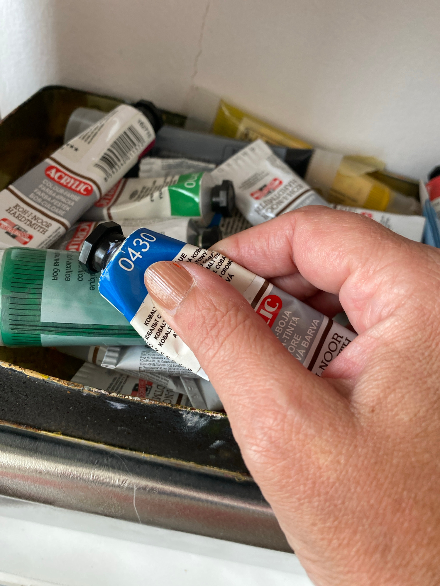 An overhead shot shows a light-skinned hand, with a polished fingernail, reaching into a container filled with numerous paint tubes. The hand is holding a blue tube of acrylic paint, seemingly in the process of selecting or retrieving it.
The container is a collection of various paint tubes, mostly appearing to be acrylics, with different colors visible such as brown, green, yellow, grey, and white. Many of the tubes belong to the brand "Koh-i-noor Hardtmuth." The specific tube being held is prominently blue with the number "0430" printed in white.
Visible text on the held tube includes "KOH-I-NOOR HARDTMUTH," "LIC," "KOBALT" (repeated in several languages including Cyrillic "КОБАЛЬТ"), and phrases like "A-TINTA" and "VÁ BARVA." Other text indicates "ROMANIA SRL" and "КОХИНОР-БЪЛГАРИЯ" ООД.
A brown tube in the foreground is also labeled "ACRYLIC" and "KOH-I-NOOR HARDTMUTH," with descriptive text such as "COLOUR FARBE," "FARBA APACKA," and "BOR-CULOARE," along with a number "162716." Other partially visible labels include "CRILIC," "Mori acrilice," "Nique," and "крилна боя." The setting appears to be an artist's workspace in Bílina, Czechia, indicated by the art supplies.