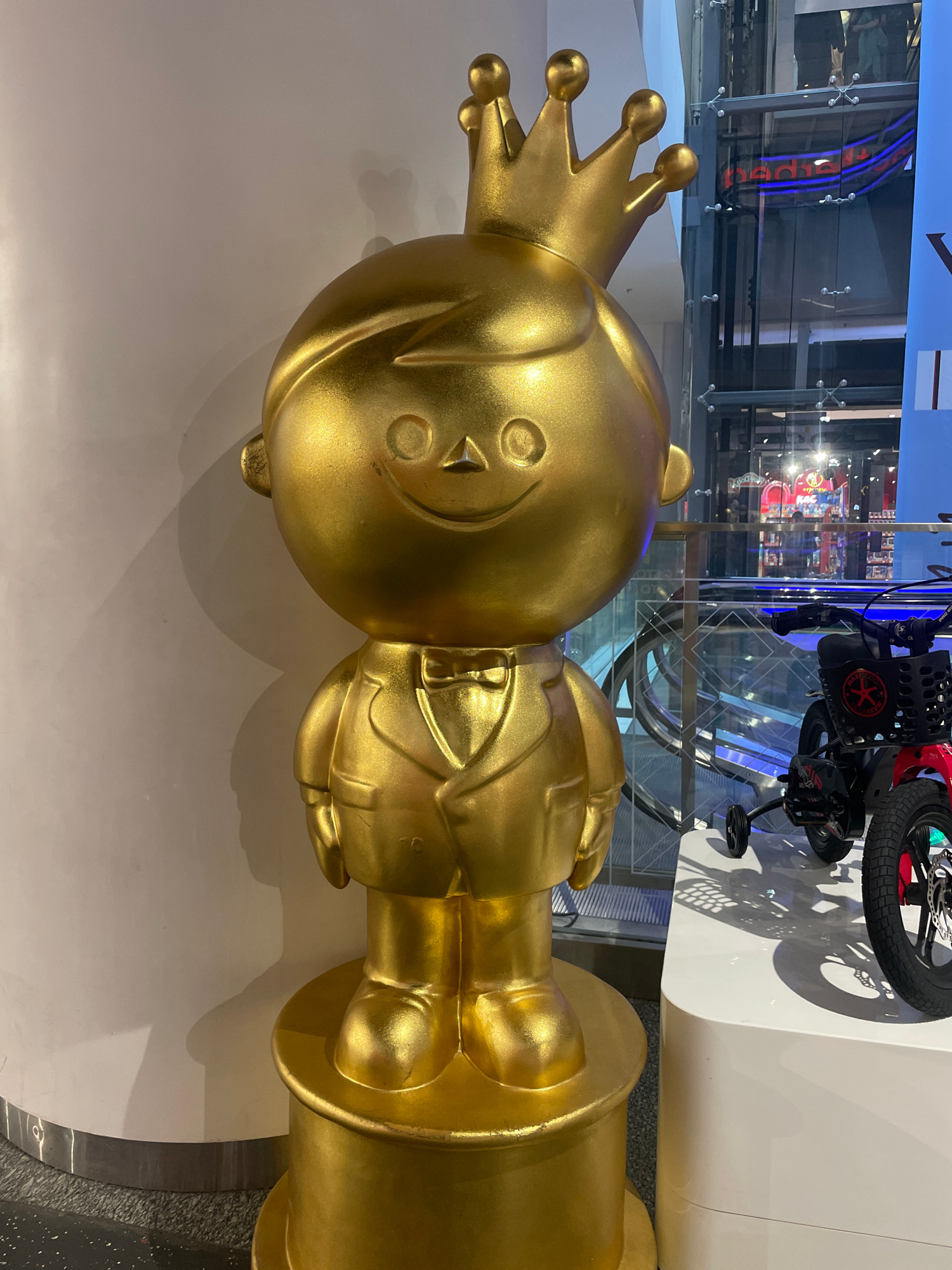 An eye-level, medium shot captures a golden, cartoonish sculpture of a young boy wearing a crown and a tuxedo, positioned in what appears to be an indoor public space. The sculpture stands on a golden circular pedestal. To the right of the sculpture, a red and black child's bicycle is displayed on a white platform. In the background, a glass elevator shaft with visible stairs and escalators suggests a mall or similar commercial building. The lighting is bright, casting shadows on the wall behind the sculpture. The overall scene suggests a retail or exhibition environment.
