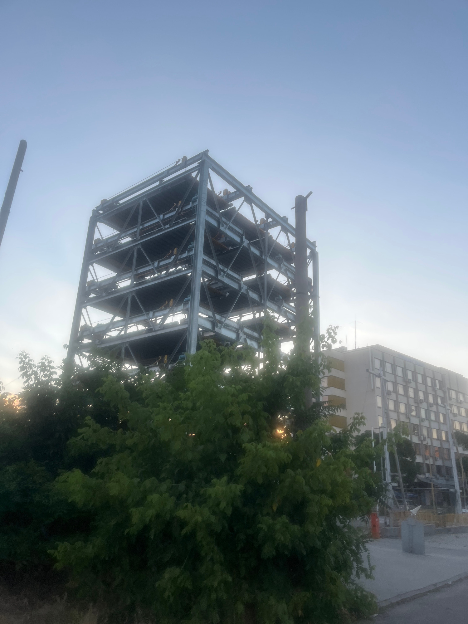 A multi-story metal frame structure, possibly a parking garage under construction or deconstruction, stands against a hazy blue sky. The structure features several horizontal platforms supported by a vertical steel framework. Green foliage is visible in the foreground, partially obscuring the lower levels of the metal structure. To the right, a modern building with a grid of windows and air conditioning units is visible. The ground in front of the building is paved, with some construction materials and barriers present. The overall impression is of an urban environment with ongoing construction or renovation.