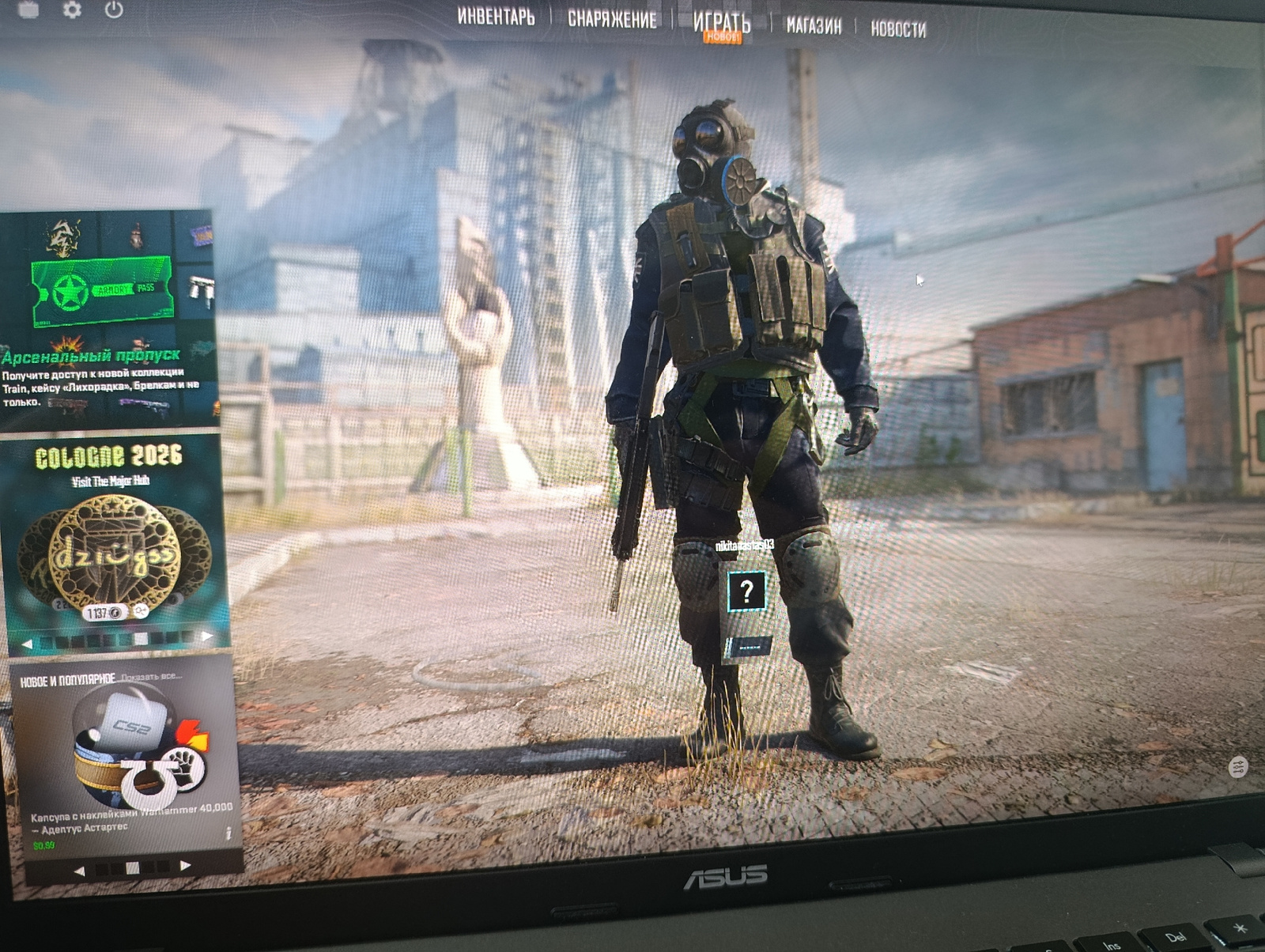 The image shows a computer screen displaying the main menu of a video game. In the foreground, a character in tactical gear, including a gas mask, vest, and combat boots, stands facing the viewer. The character's name tag reads "nikitavasts03". To the left of the character, a menu panel is visible with various game-related information. This panel displays options such as "Armoury Pass," "Cologne 2026," and "New and Popular." The background depicts an urban environment with dilapidated buildings, suggesting a post-apocalyptic or industrial setting. The overall scene is a digital rendering, characteristic of a video game interface. There is no indication of the city of Rezina, Moldova, or any specific real-world location within the image.